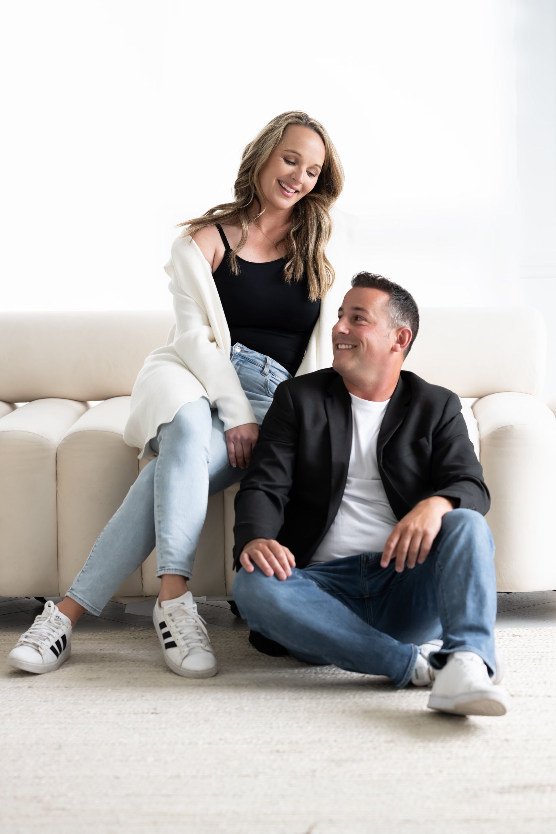 An informal and cozy portrait of a smiling couple seated on a white couch, conveying a relaxed yet professional ambiance.