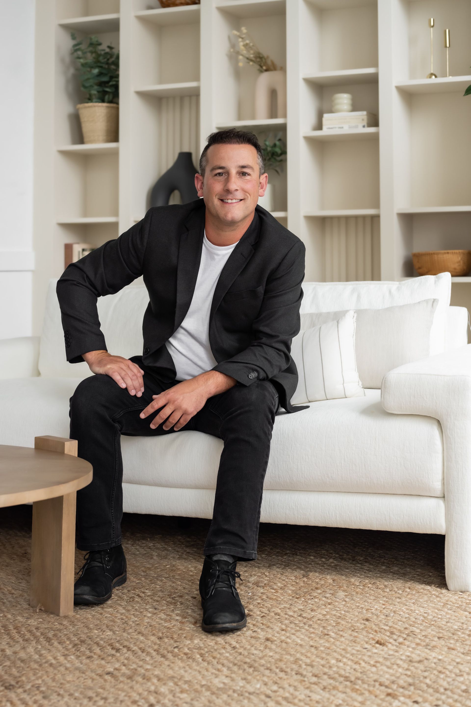 A man in a smart black jacket and jeans sits casually on a white sofa, exuding confidence and approachability, set against a chic and organized bookshelf backdrop.
