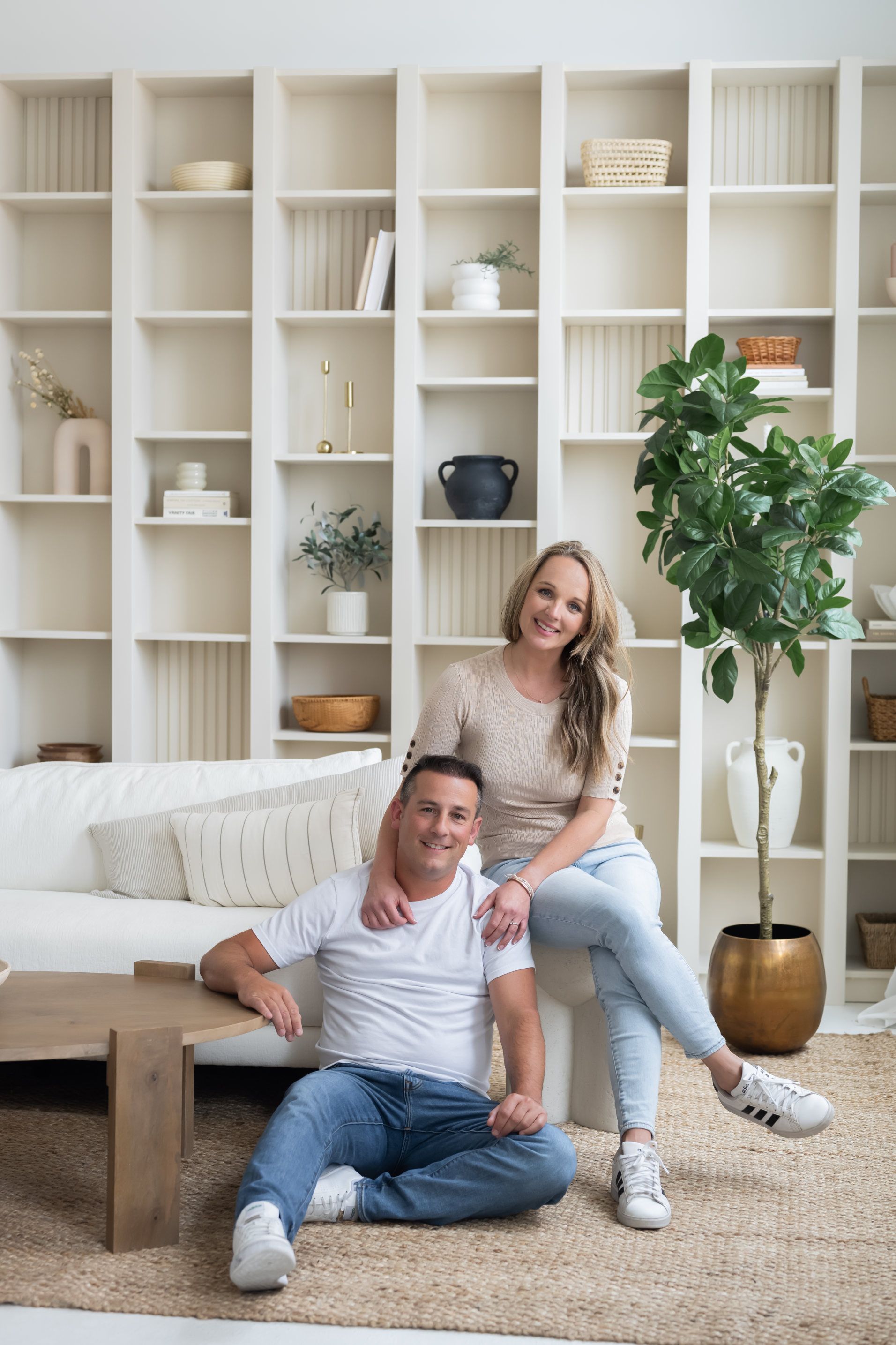 A smiling couple in casual attire sits comfortably in a living room, exuding a relaxed yet stylish vibe, with a neatly arranged bookshelf and lush greenery enhancing the homely atmosphere.