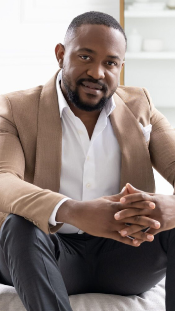A man in a suit is sitting on a couch with his hands folded.