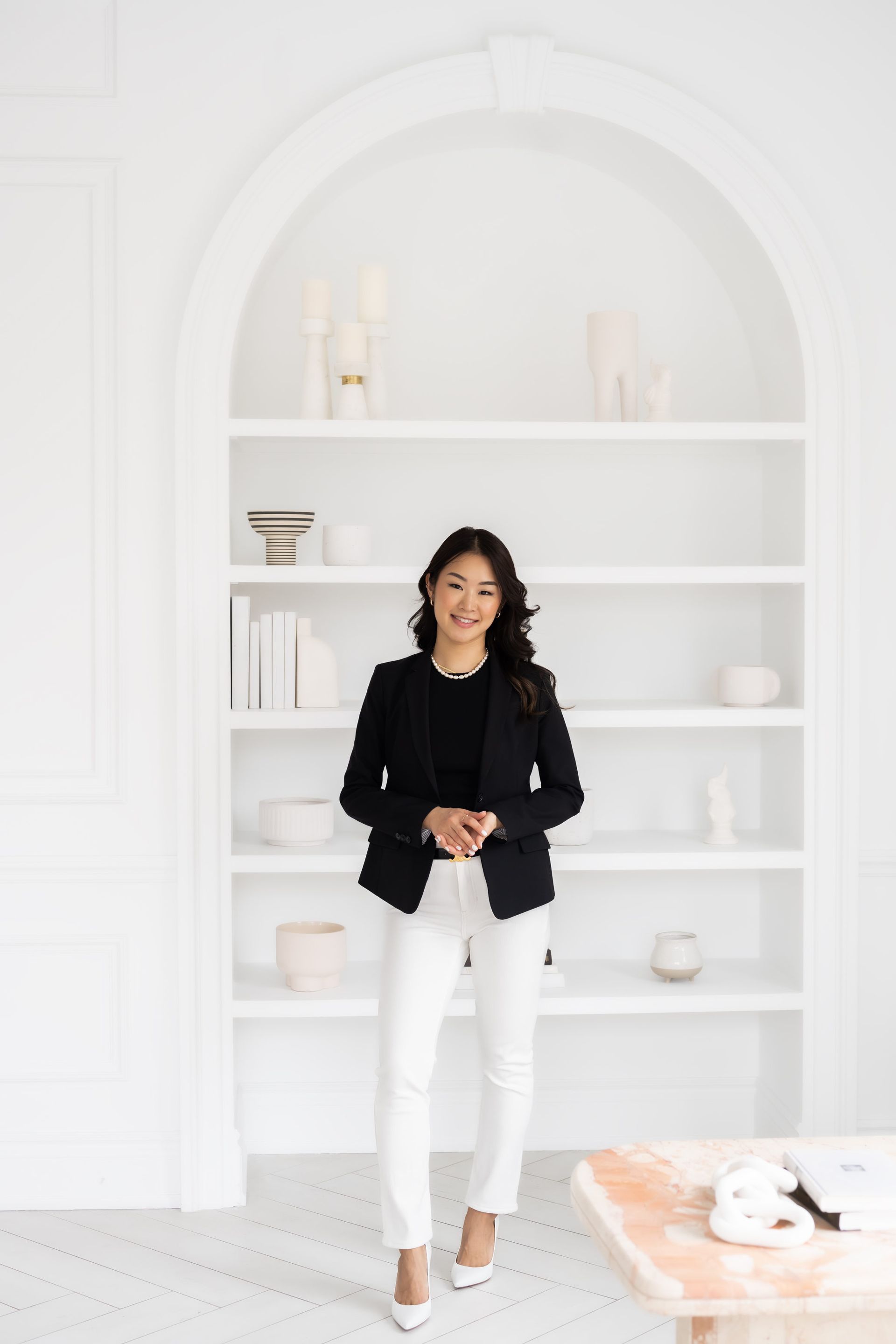 a woman in a black jacket and white pants standing in front of a white shelf.