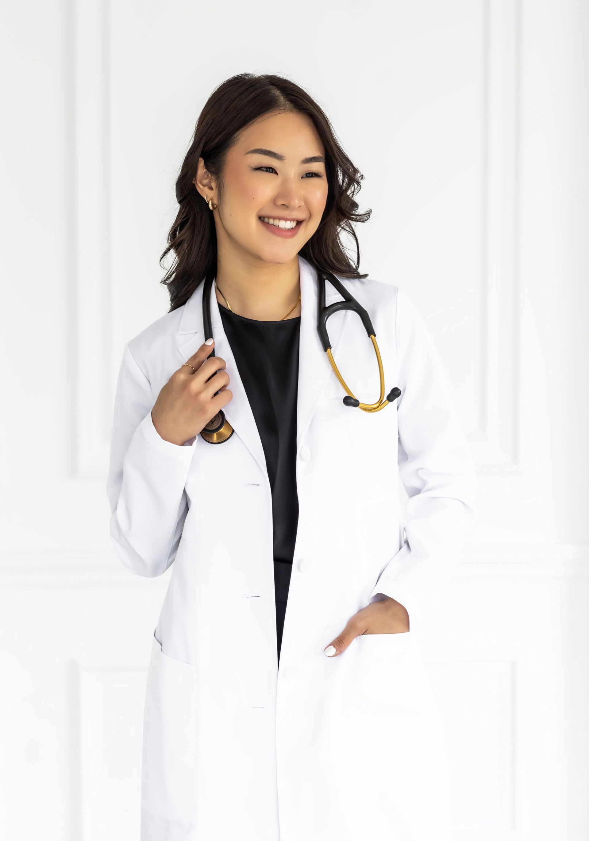 Smiling medical professional in a white jacket and black top, with a stethoscope around her neck, standing confidently in a bright white room.