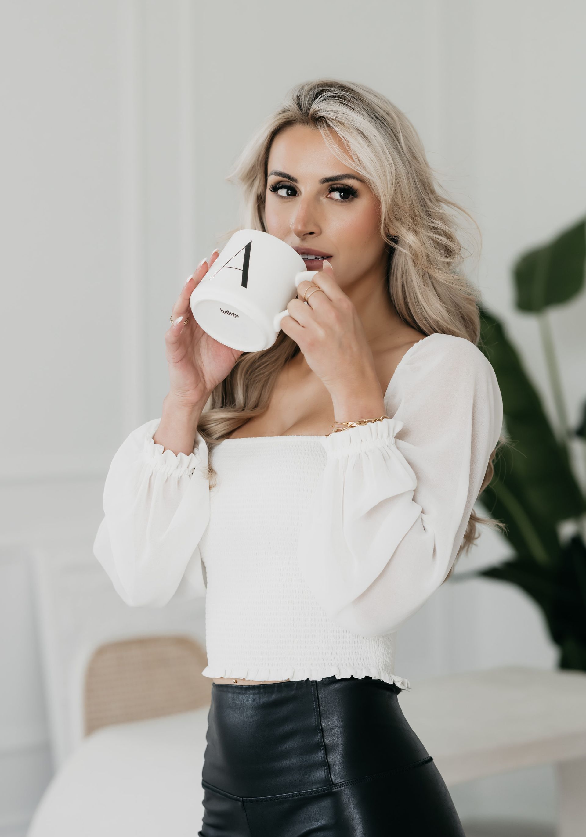 Woman in black leather skirt and white blouse poised to sip from a large coffee mug, with a focused gaze directed off to the side in a personal branding photography setting.
