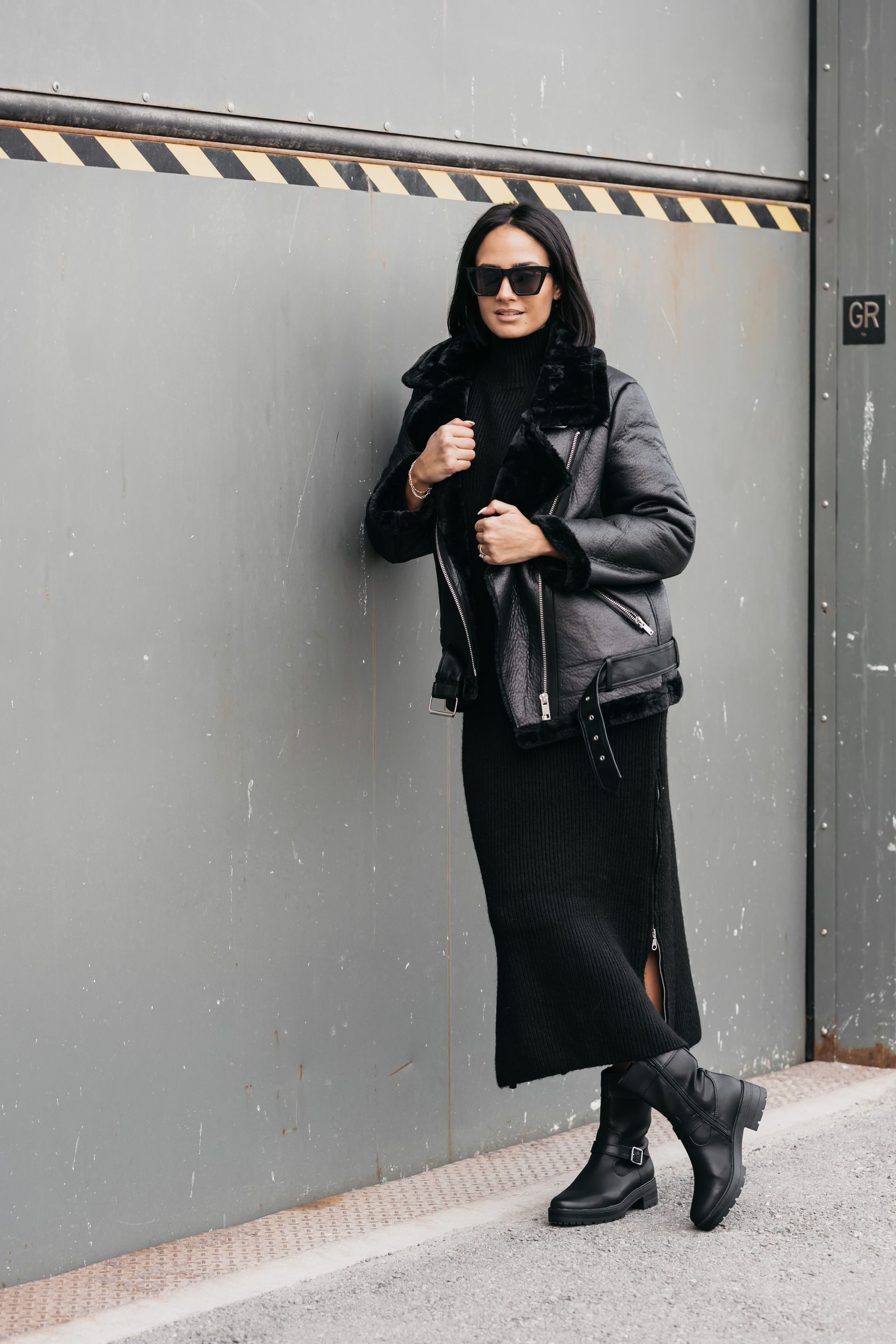 Woman in a long black skirt, motorcycle-style leather coat, ankle-high boots, and dark sunglasses, leaning confidently against a closed freight elevator door for her personal branding photos.