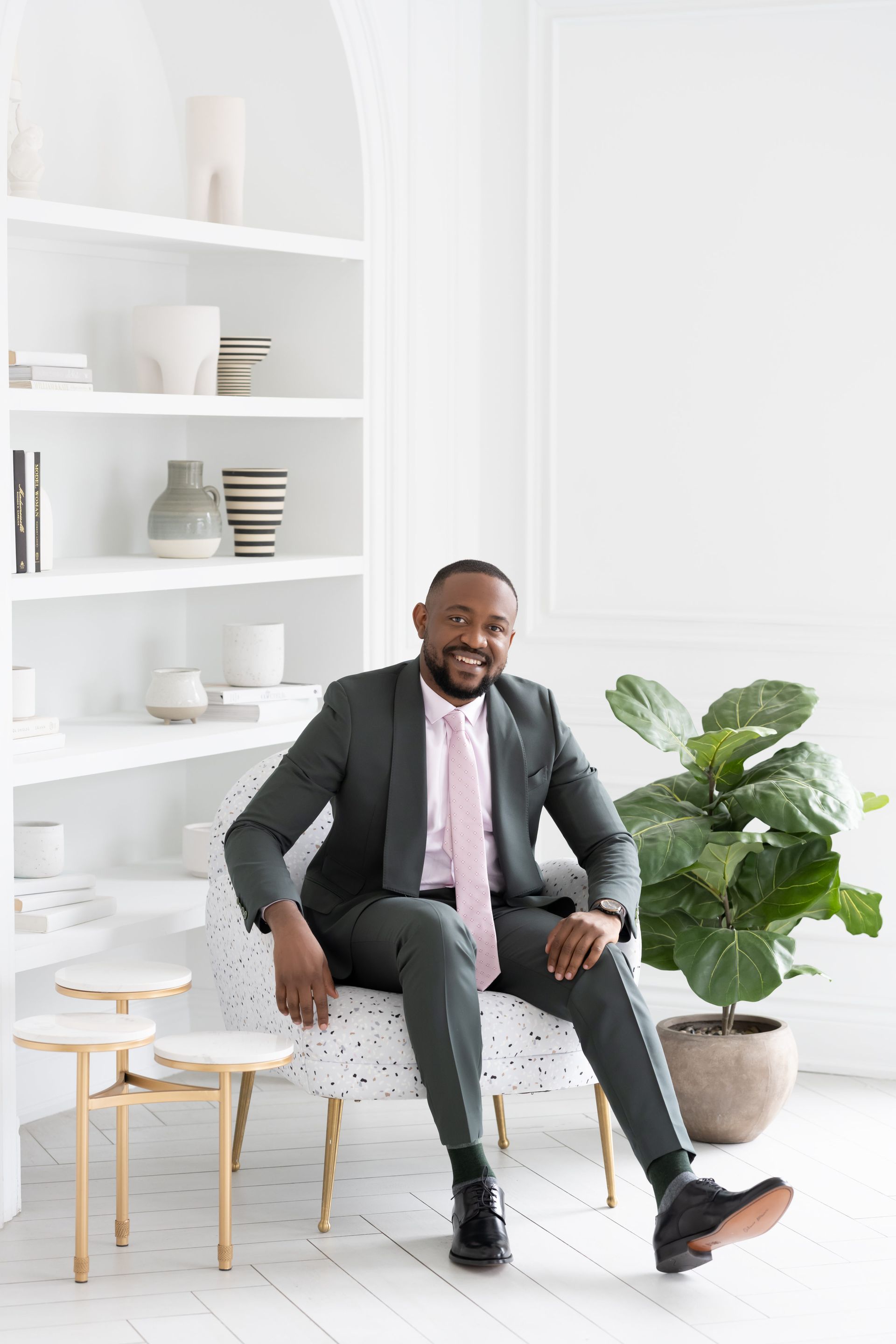 Engaging man in suit and tie seated in a white room with minimalistic décor including a plant and built-in cabinet. He offers a welcoming smile while leaning forward in his chair for his personal brand photography session