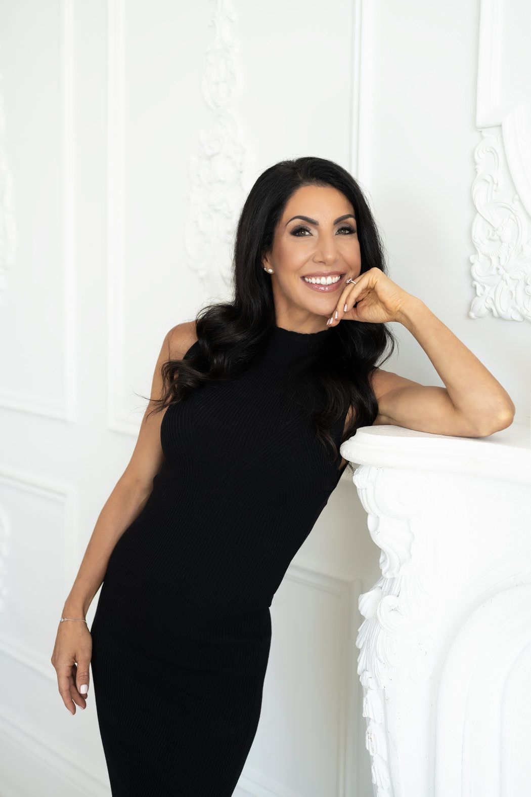 Woman in a contrasting black sleeveless dress, smiling engagingly while resting her elbow on an all-white fireplace mantle, creating a captivating personal branding photography scene.