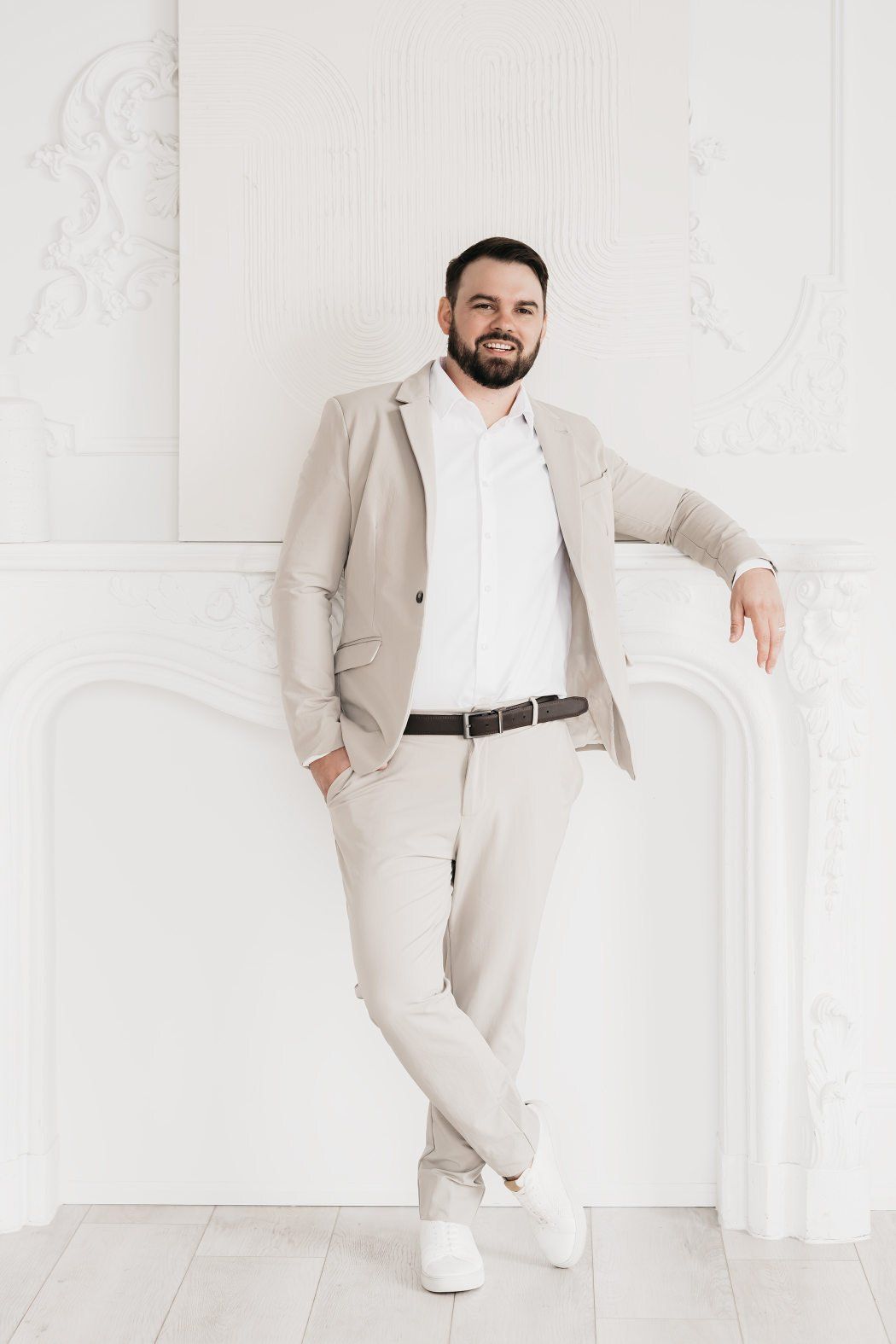 A man wearing a suit stands against a clean white wall, exuding professionalism.
