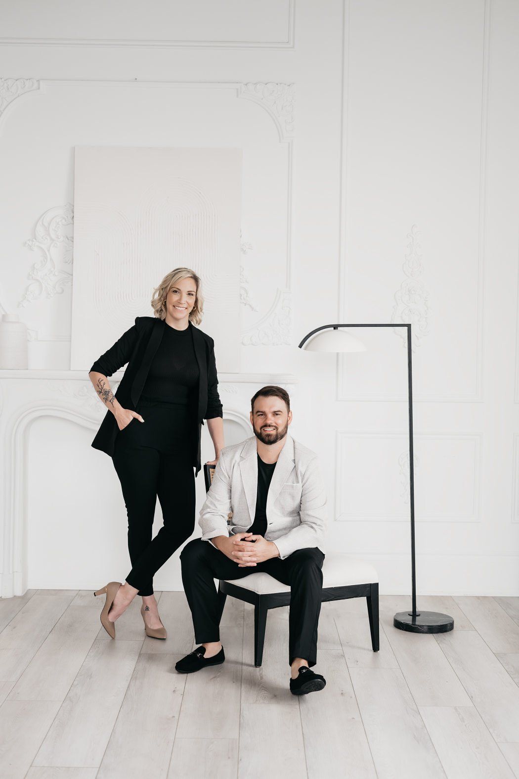 A couple sits together on a chair in front of a plain white wall, smiling.