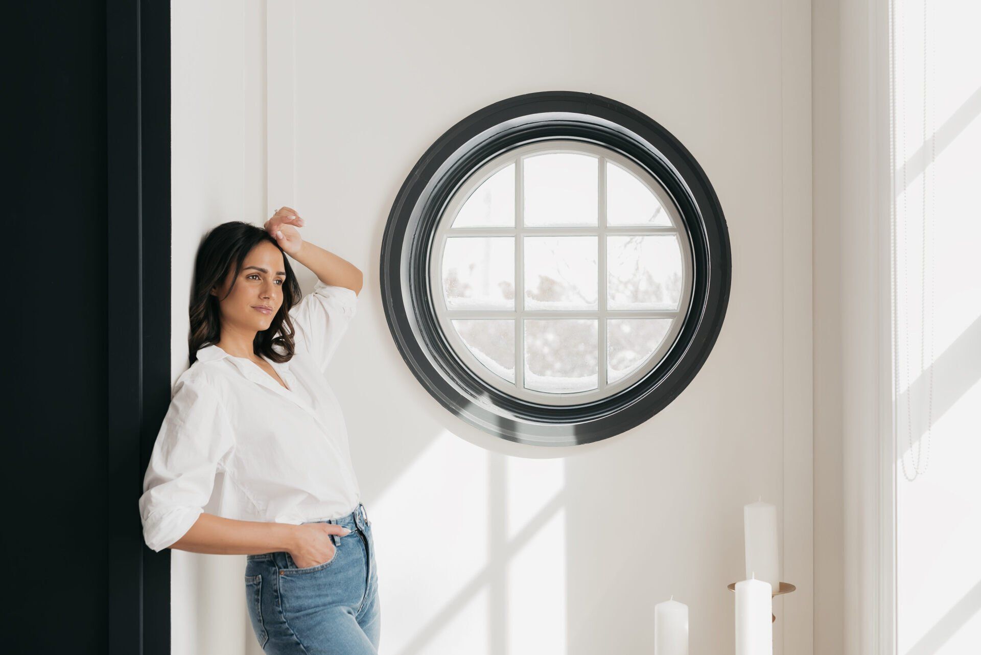 A young woman wearing a crisp white shirt and jeans posing beside a round window with natural lighting in the hallway of a manor