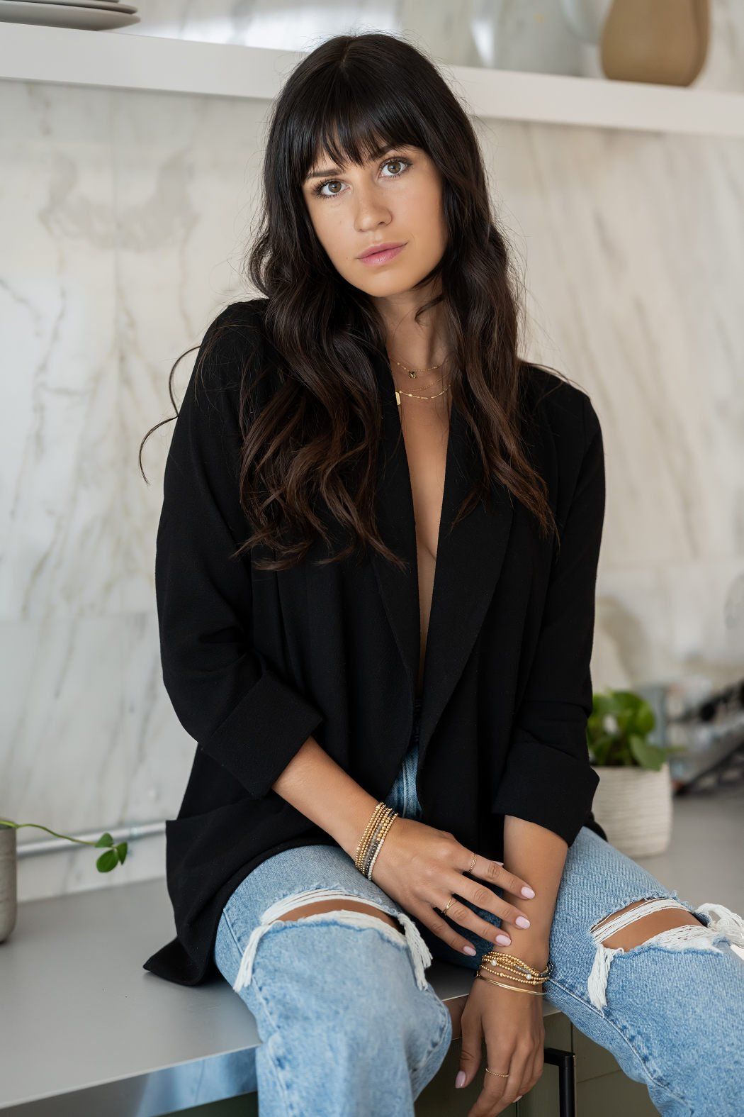 A young woman with wavy hair and heavy bangs wearing distressed jeans and a black blazer sitting on a kitchen counter with marble walls