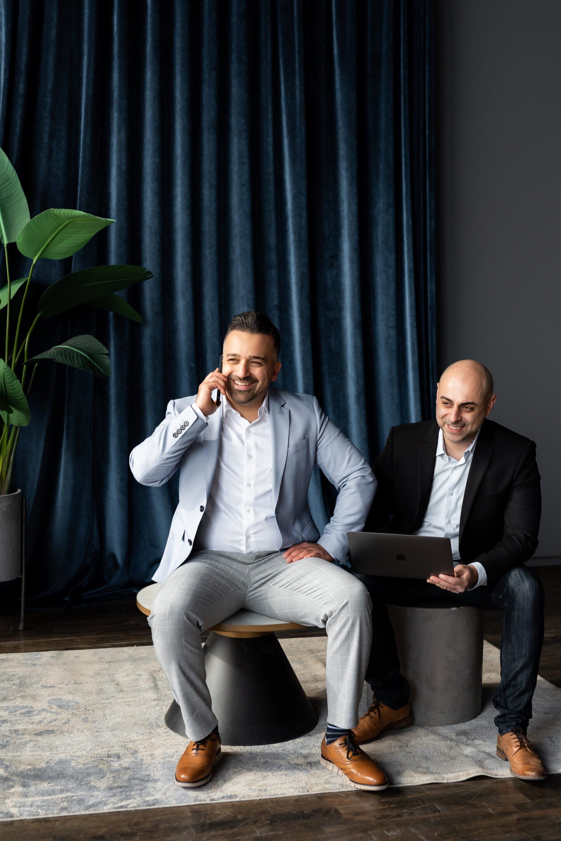 two realtors, one is smiling while in phone call, the other is smiling while using his laptop
