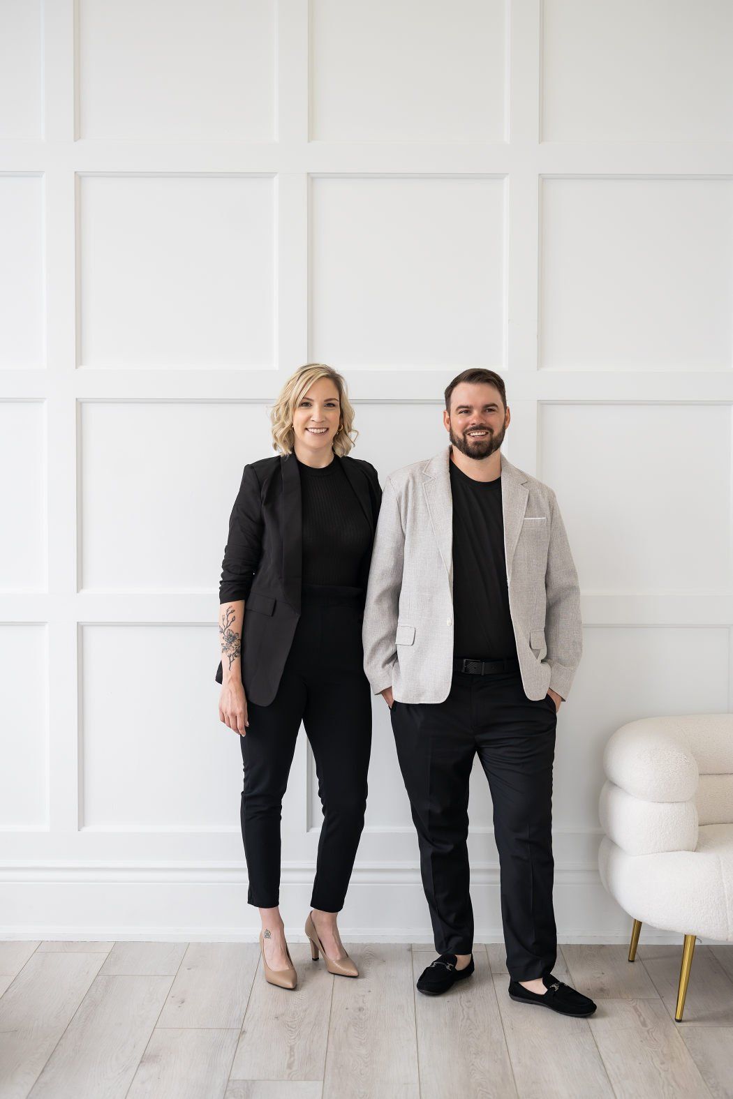 A couple wearing smart casual outfits while standing in front of a white wall with square embossed details