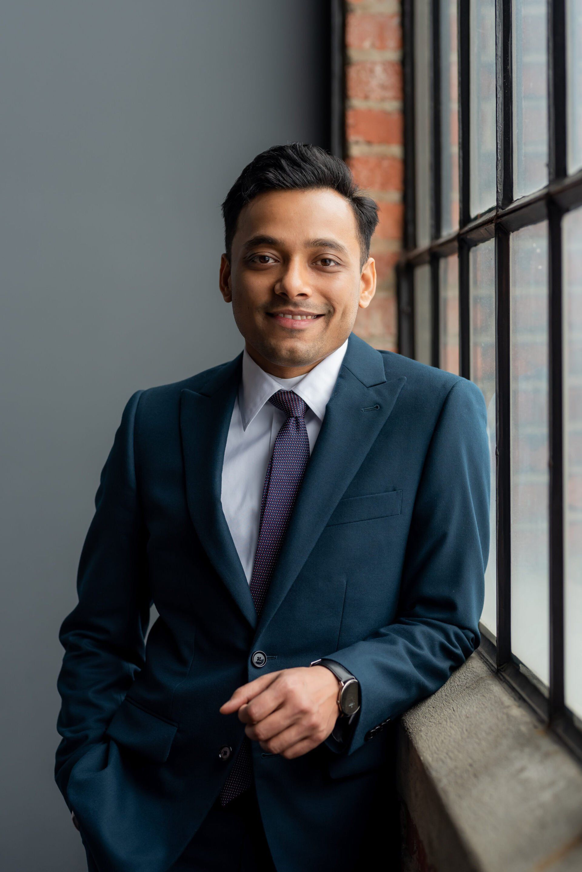 Personal branding photo of a man in a navy blue suit while leaning beside a window in an industrial style office