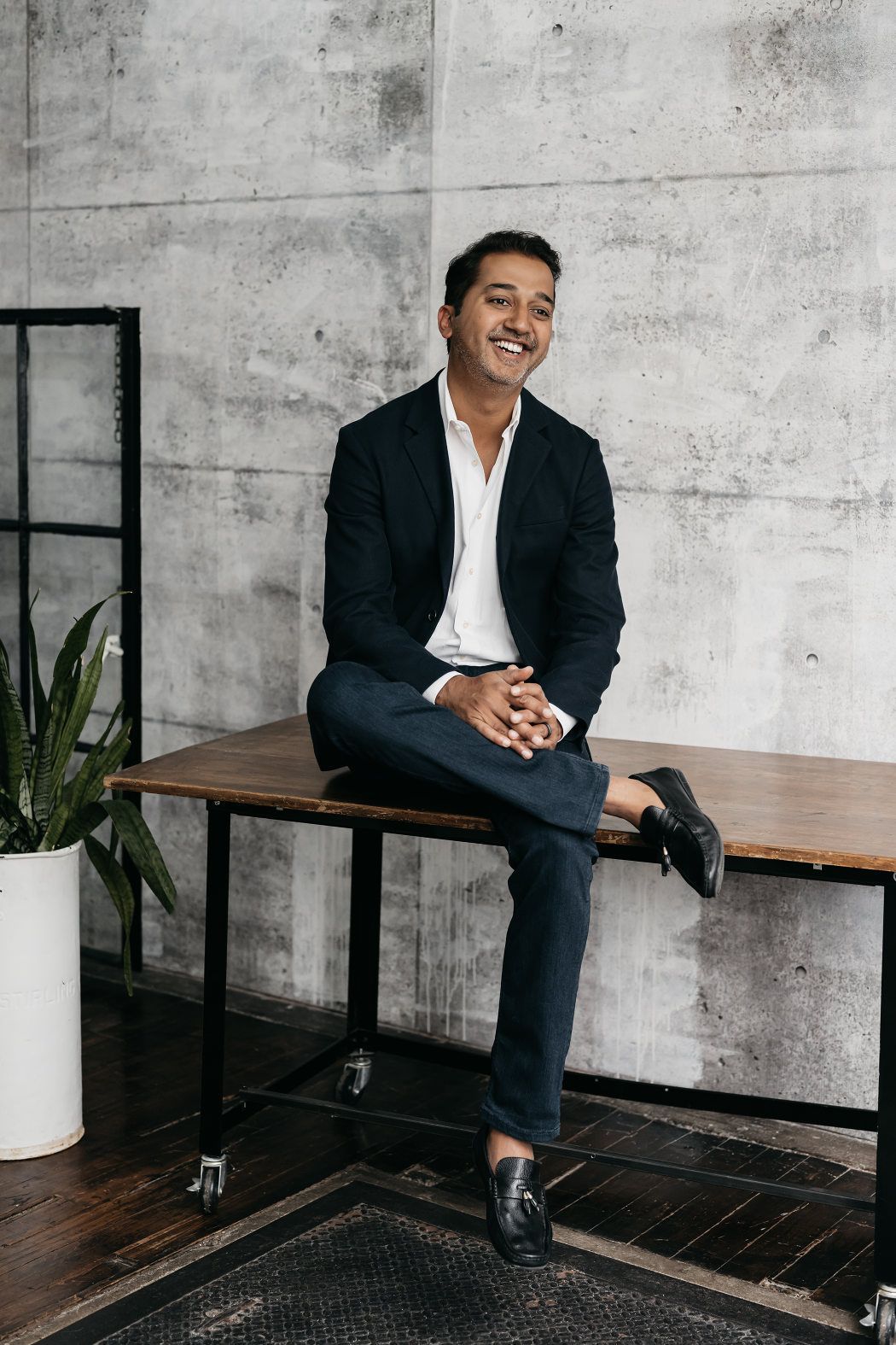 Man smiling during a personal brand photography session, seated on a table in a loft-style room with exposed concrete walls and aged wooden floors.