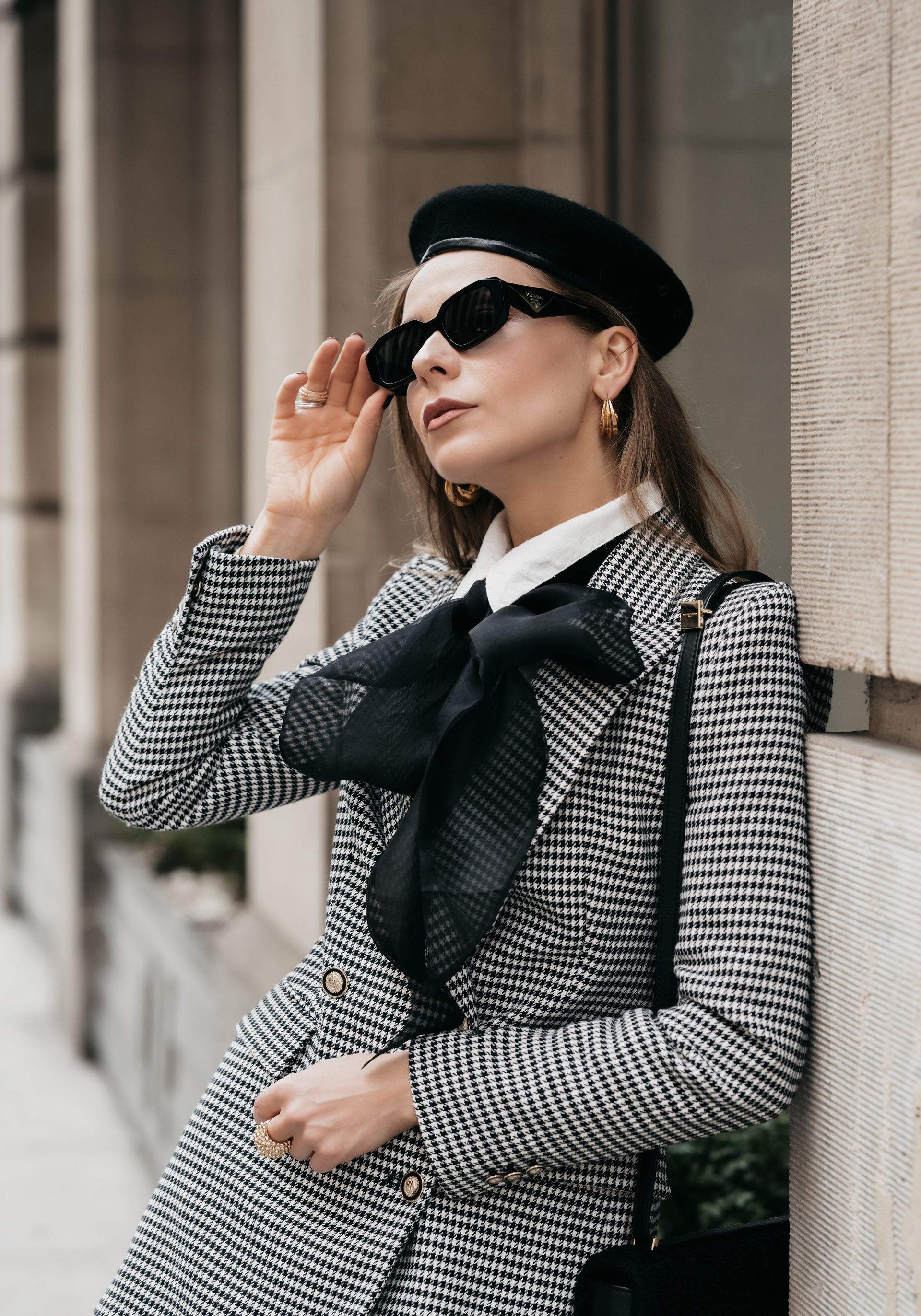 Woman in houndstooth coat, black beret, and ribbon, adjusting sunglasses against a brick backdrop for personal branding photos.