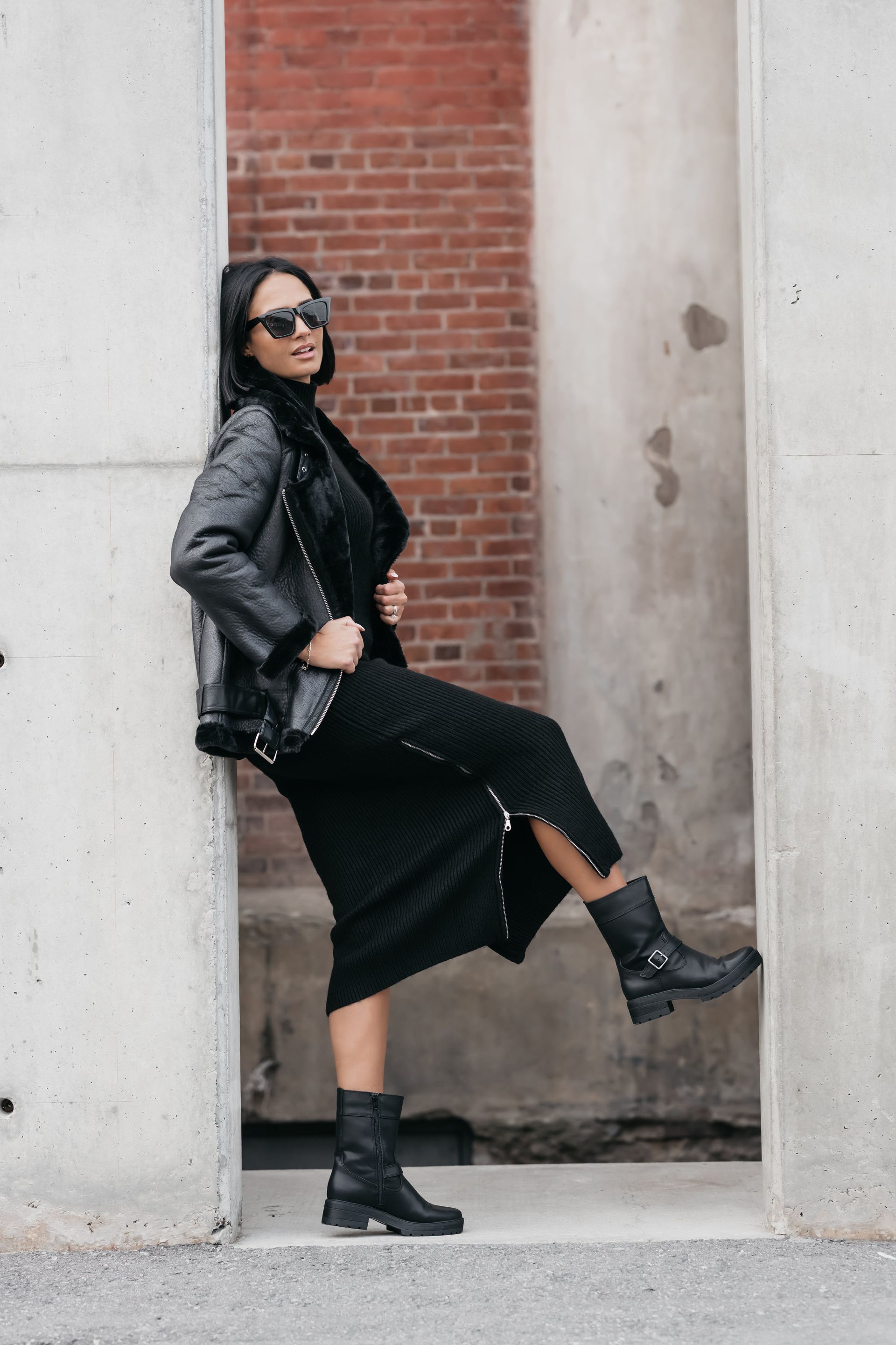 Woman in black leather jacket, skirt, and calf-high boots striking a dynamic pose between two pillars for her personal branding photo.