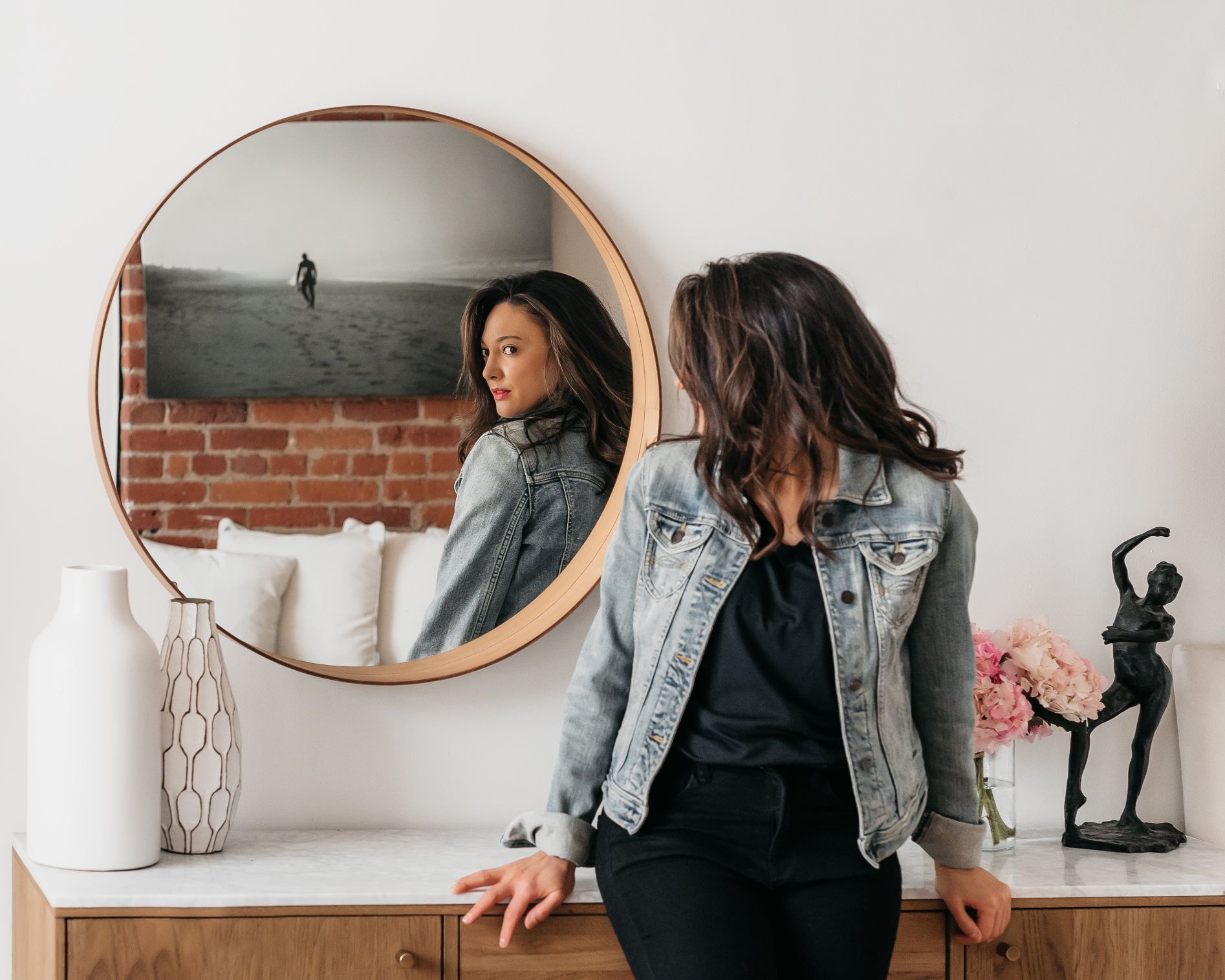 A woman is standing in front of a round mirror looking at her reflection.