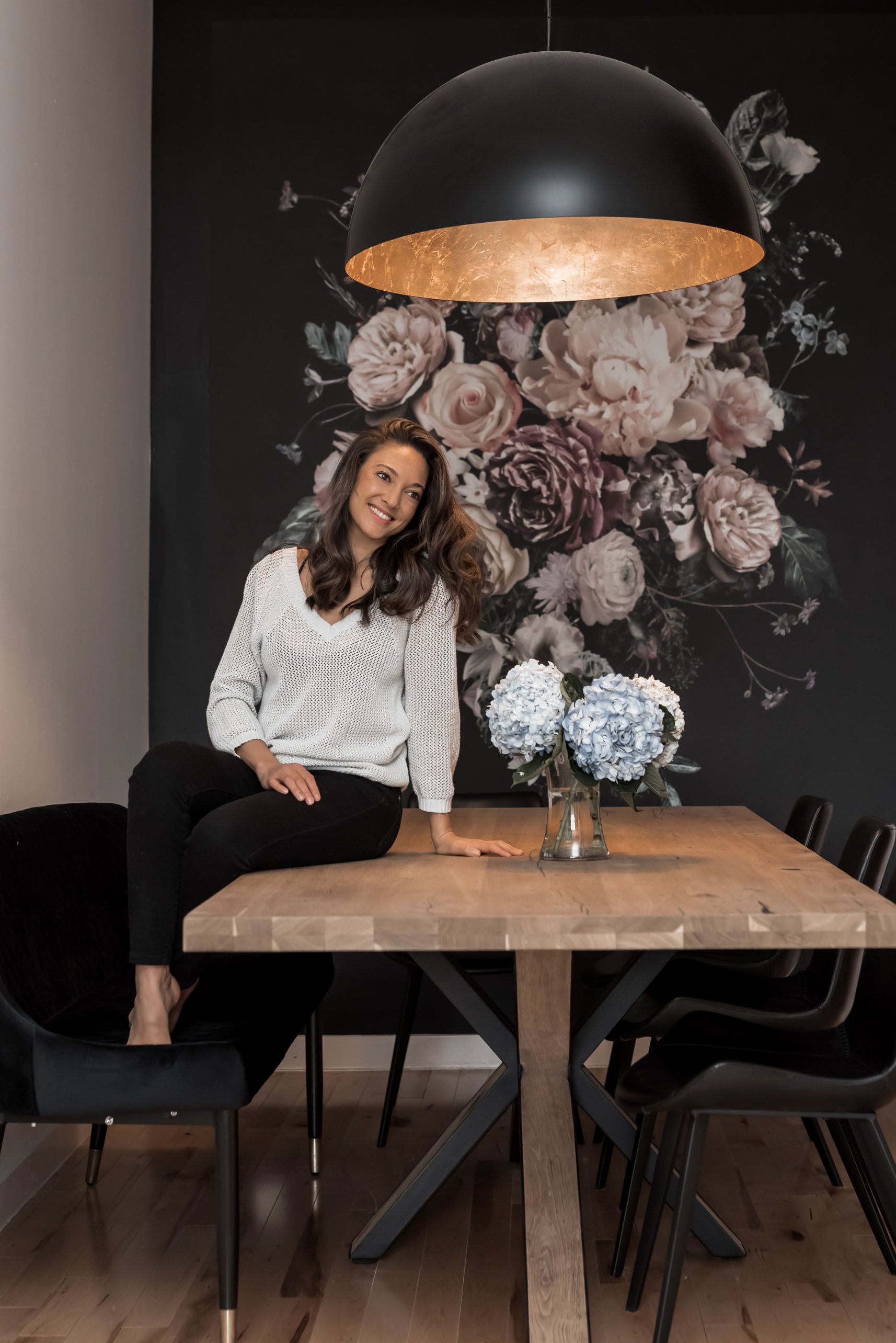 A woman is sitting at a table in front of a floral wall mural.