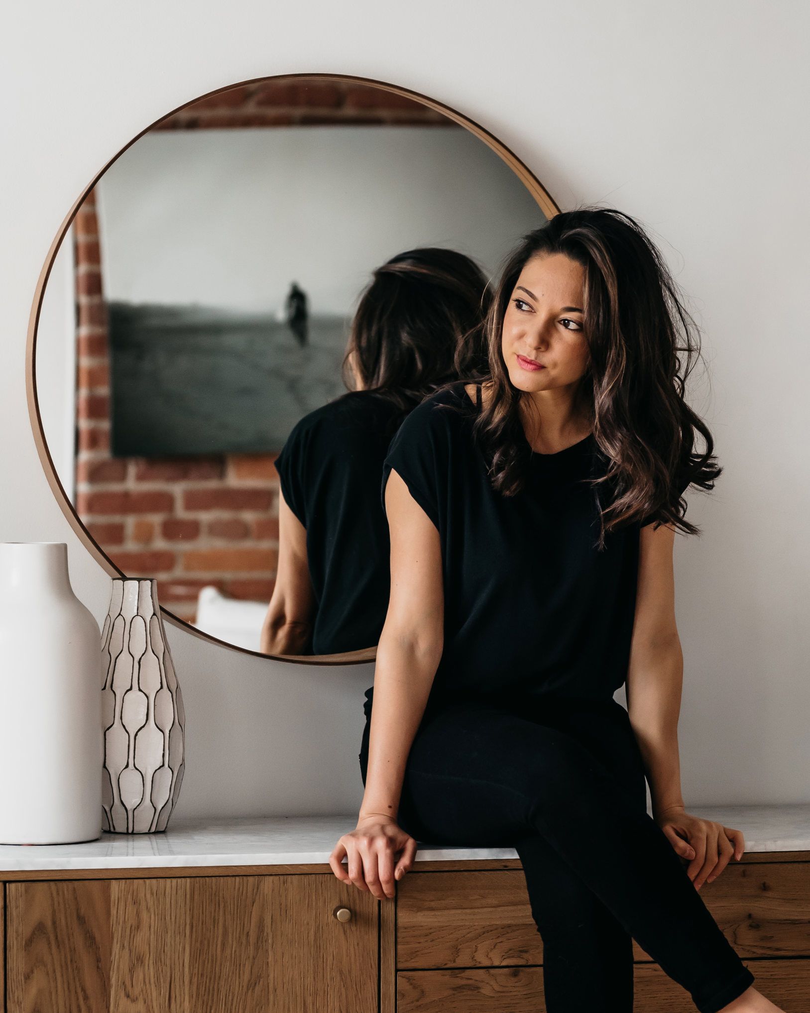 A woman is sitting on a dresser in front of a mirror.