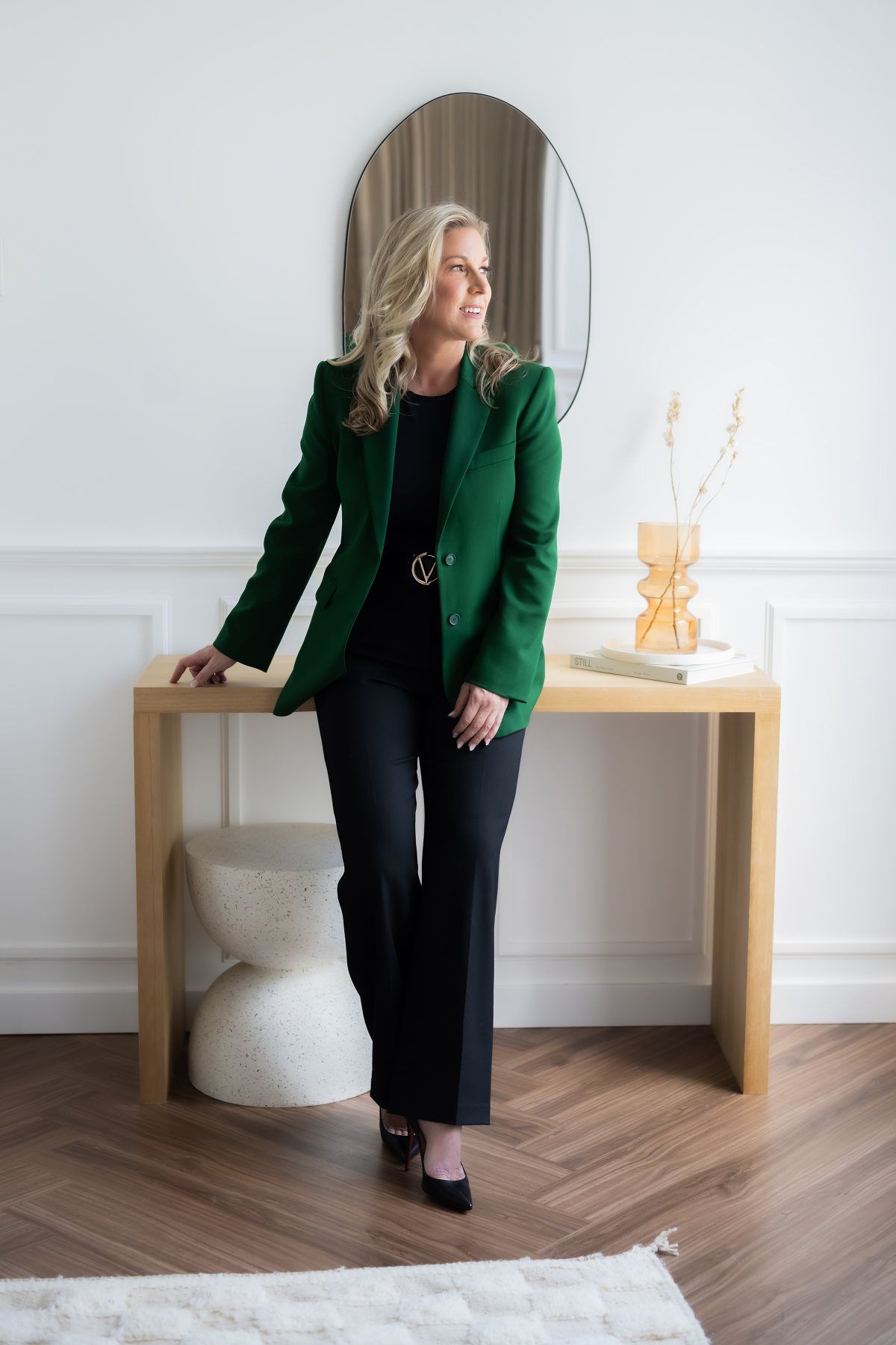 A woman in a green jacket and black pants is leaning on a wooden table.
