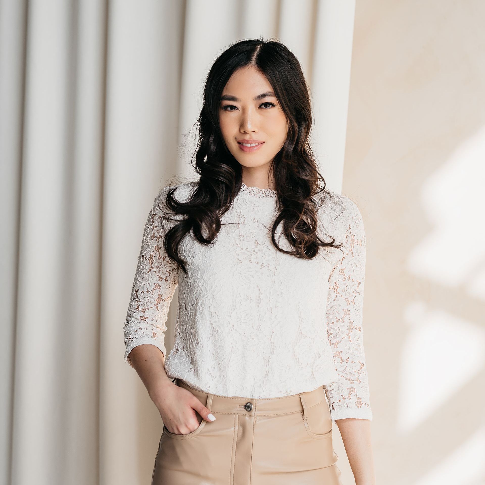 A woman wearing a white lace top and khaki pants is standing in front of a white curtain.