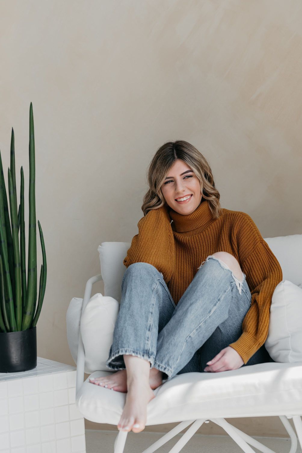 A lifestyle-themed personal branding photoshoot features a woman lounging in a cozy mustard turtleneck and distressed jeans, her joyful smile and casual pose exuding a sense of wellness and relaxed confidence.
