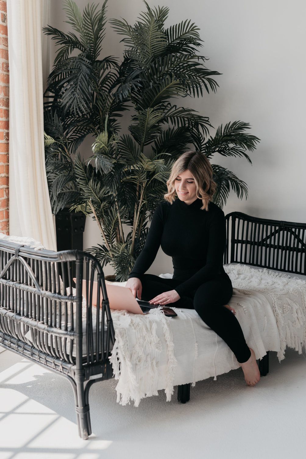 An entrepreneur sits comfortably on a white, fringed daybed, working on her laptop with a relaxed posture.