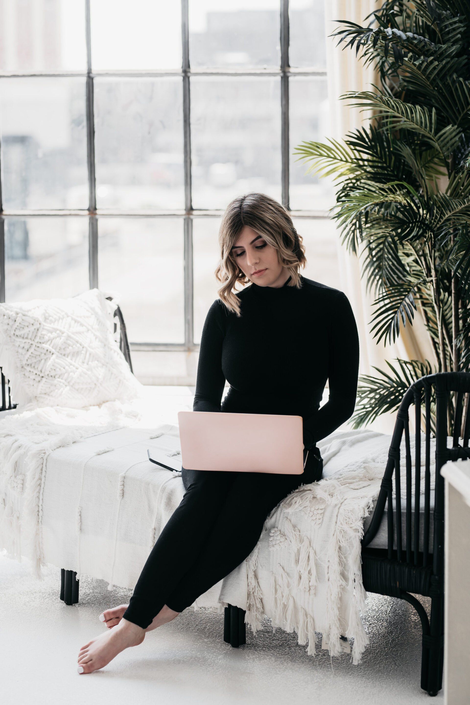 woman dressed in black pants and top, sitting on the side of a bed in an all-white room while working on a laptop
