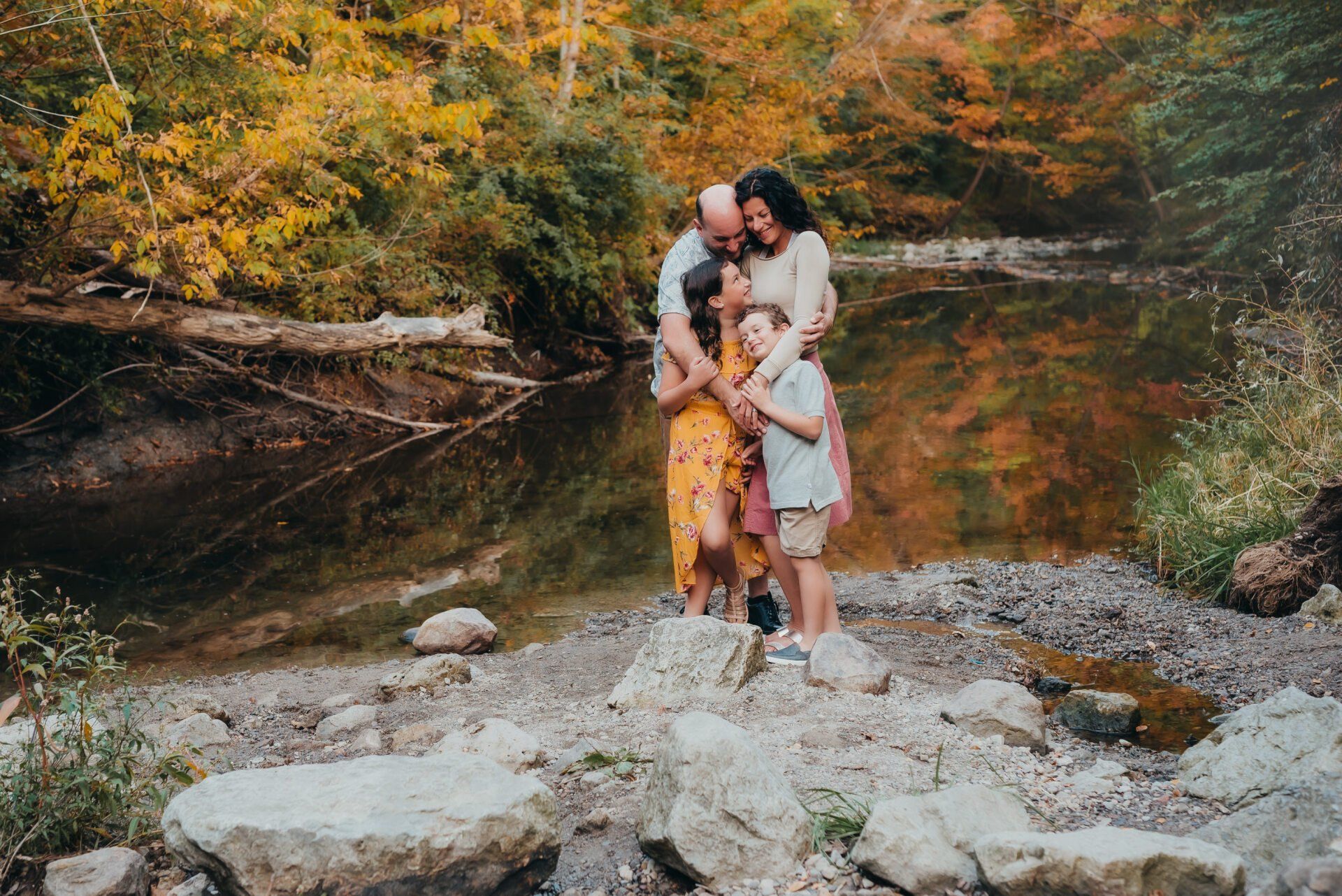 Mother, father, daughter and son holding on to each other while standing on edge of creek for fall family photoshoot