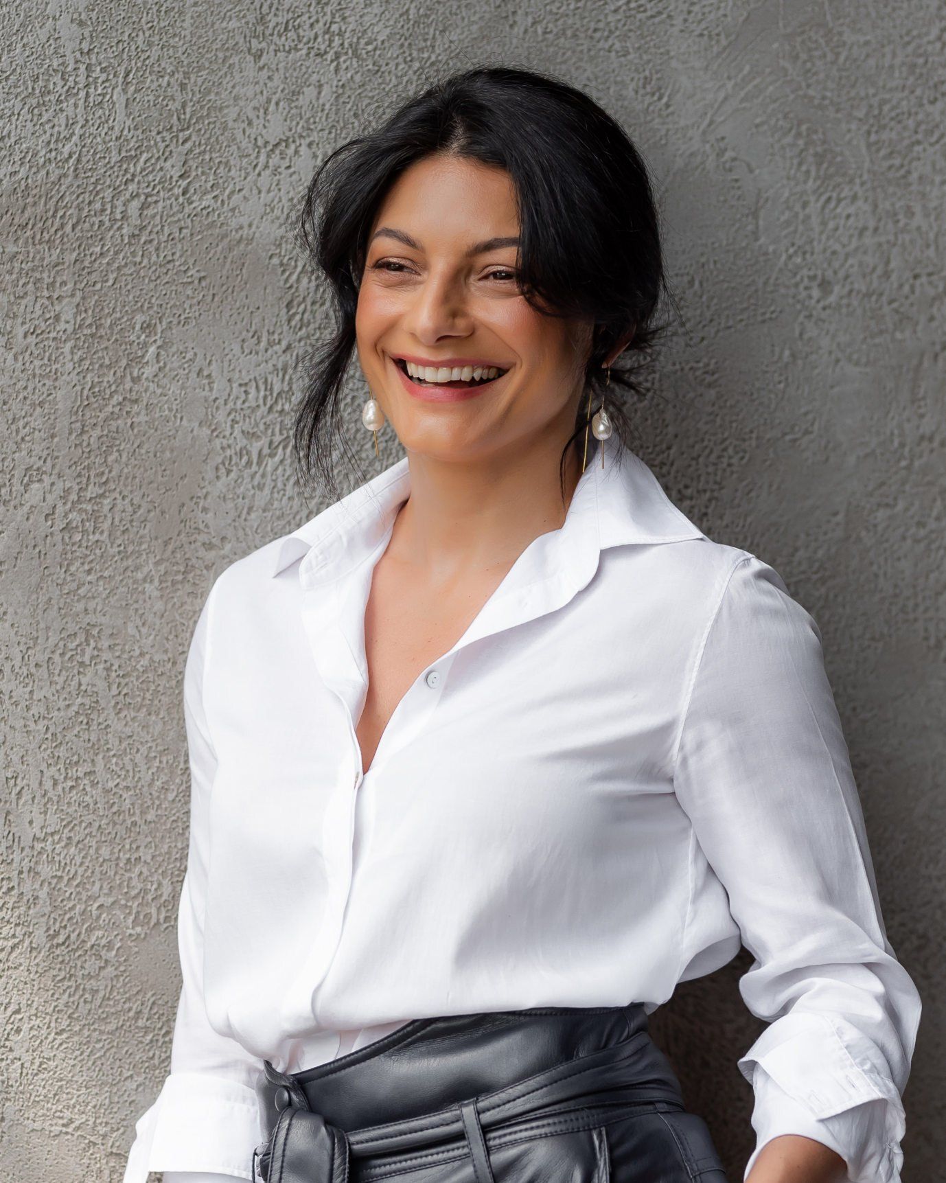 A woman in a white blouse is laughing while standing against a stucco wall.