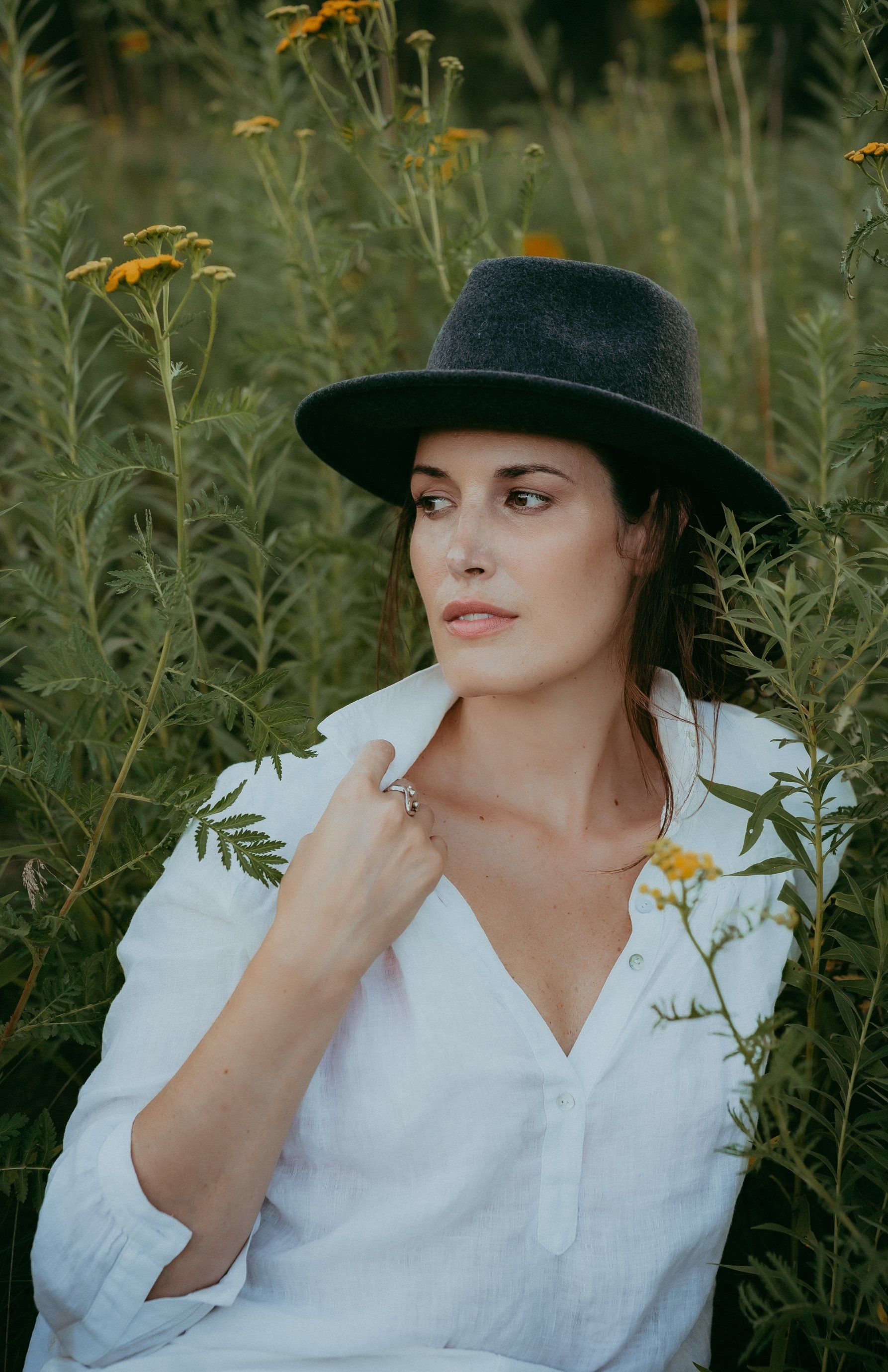 Personal branding photo of woman wearing a black hat and white blouse sitting in field of tall grasses and wild flowers