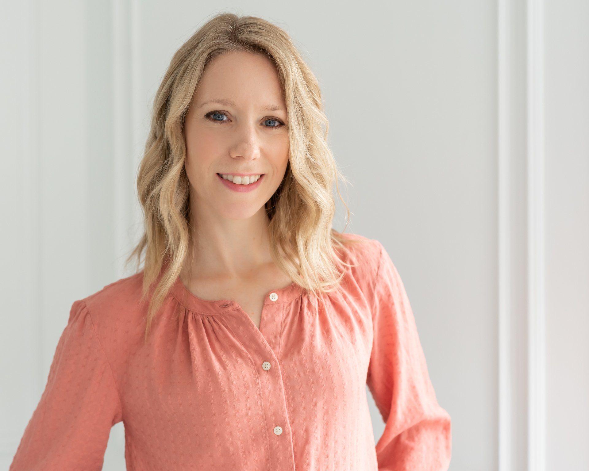 a photograph of a blonde-haired woman wearing peach top with a light background