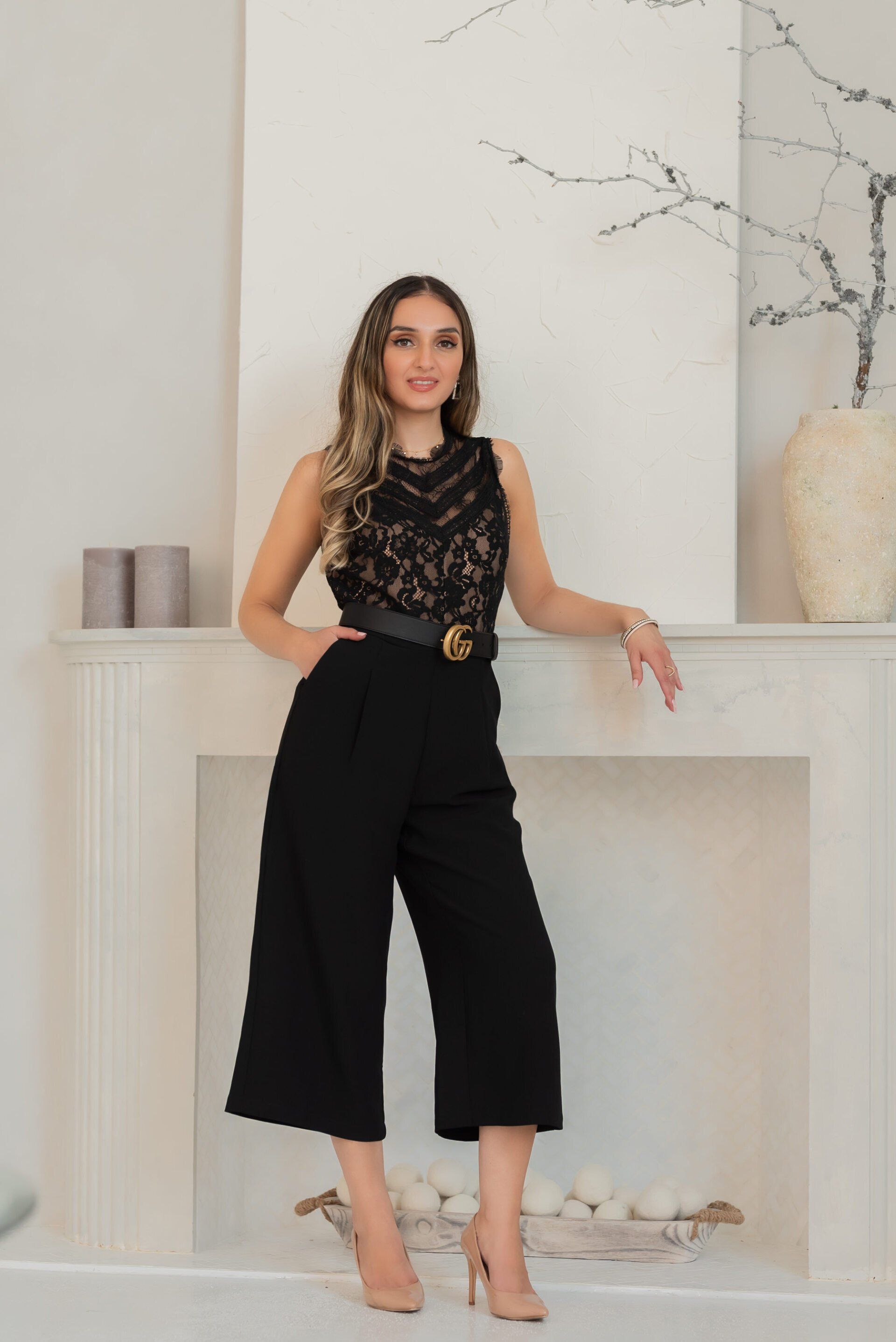 A confident young woman dressed in black pants and a black top stands against a white fireplace for a personal branding photo.