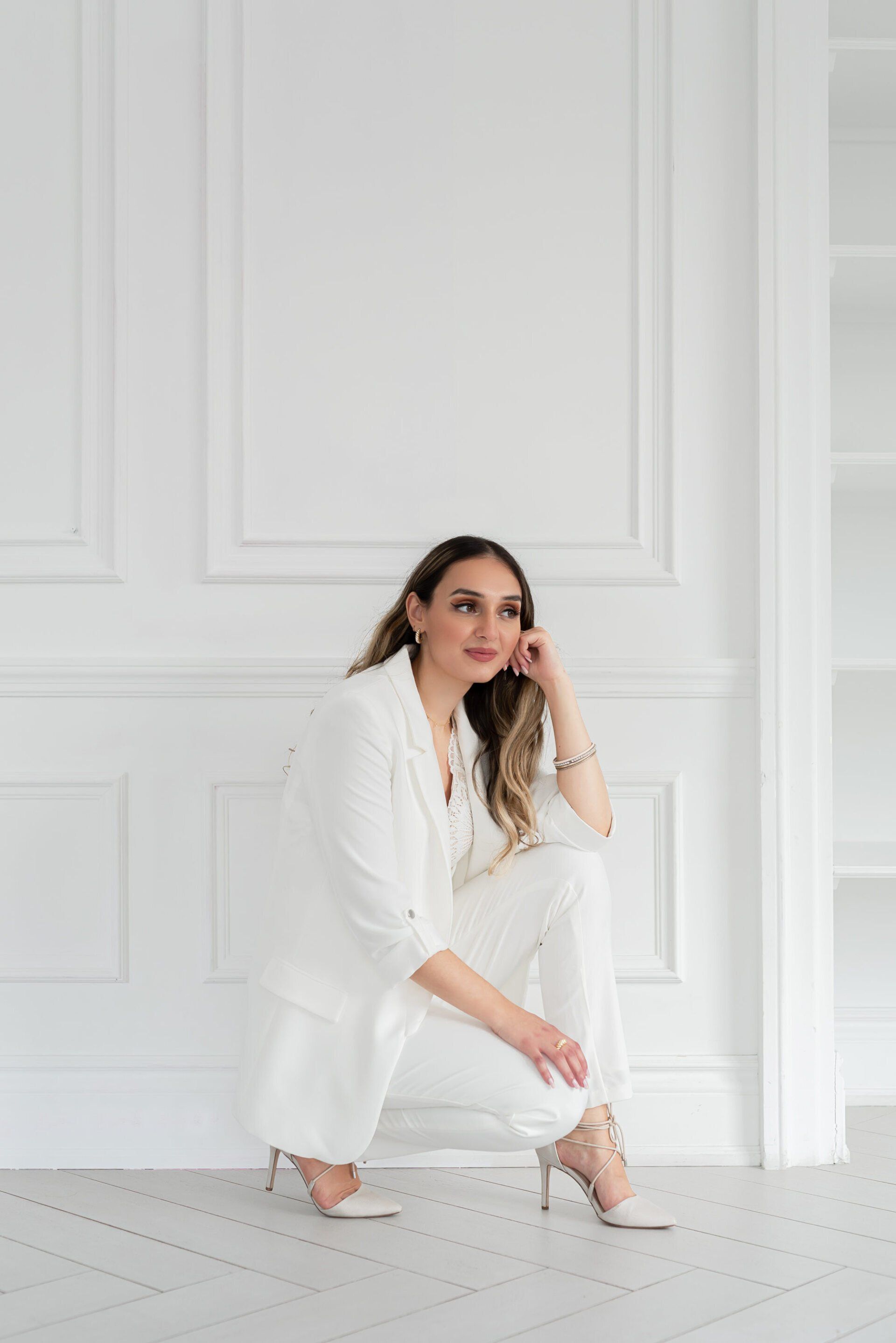 A young woman dressed in white crouches down in front of an all white wall.