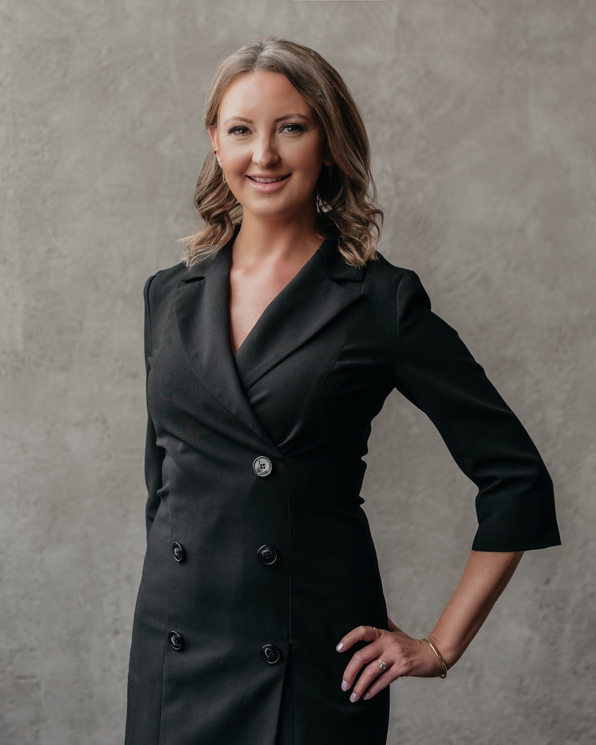 Woman with wavy brown hair in a black double-breasted blazer, standing against a gray background.