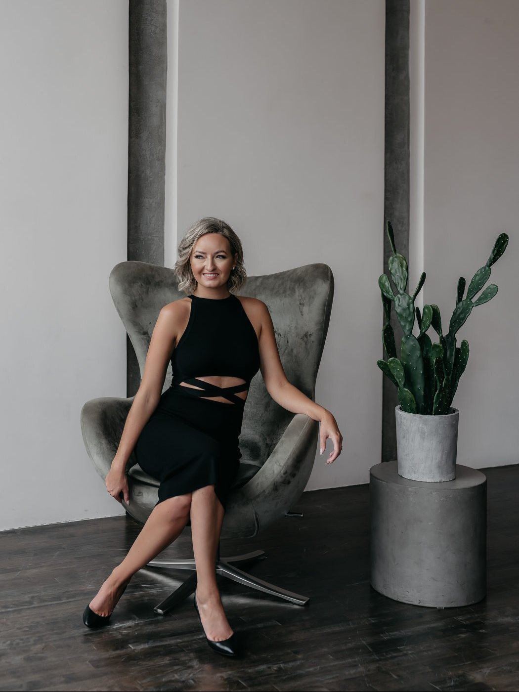 A woman in a black dress is sitting in a chair next to a plant.