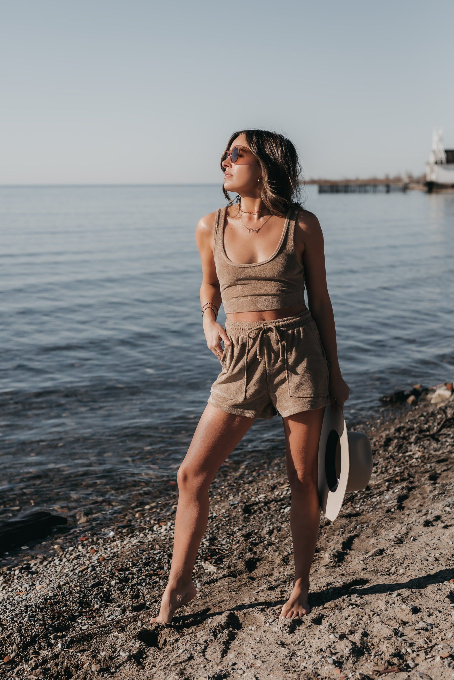 a woman posing on the shore while holding a taupe hat