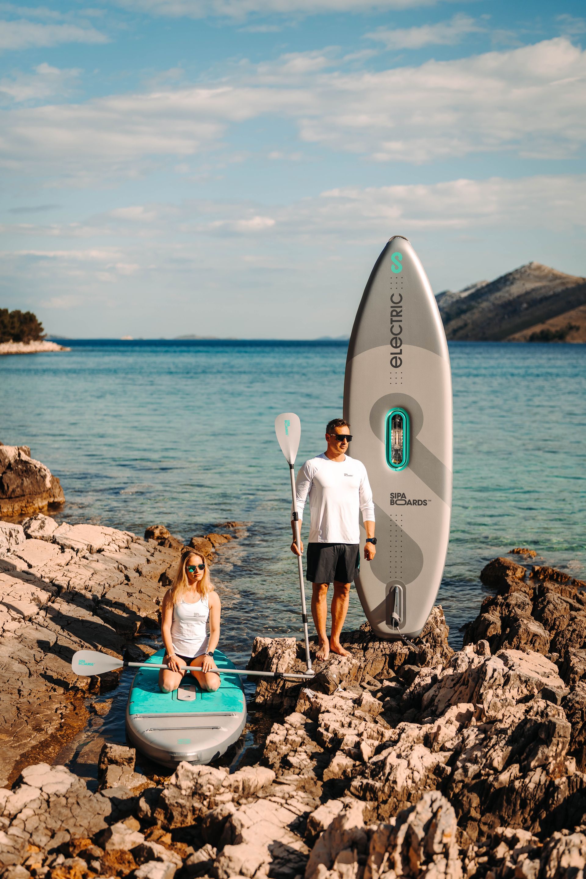 A man and a woman are standing on rocks next to a paddle board.