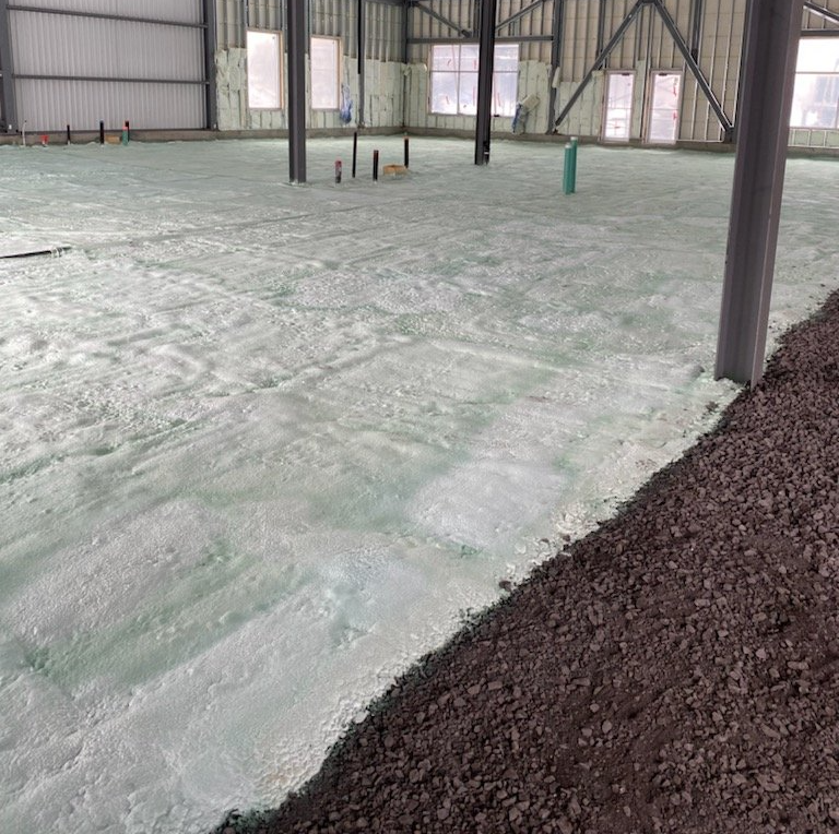 Vue intérieure d'un bâtiment avec isolation en mousse projetée au sol. Des graviers foncés bordent le sol.