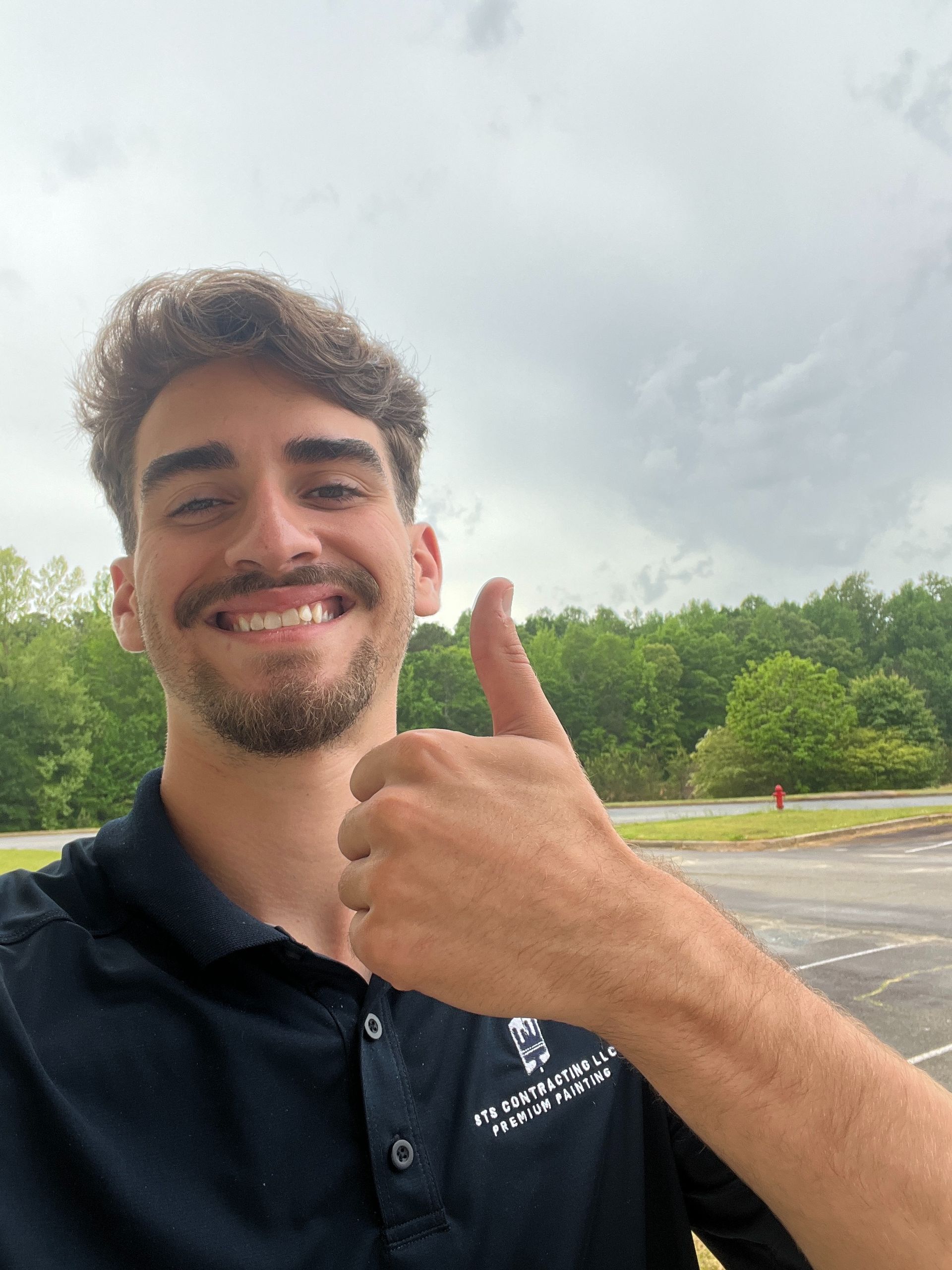 Man with curly brown hair and a beard gives a thumbs up outside. Cloudy sky and trees in background.