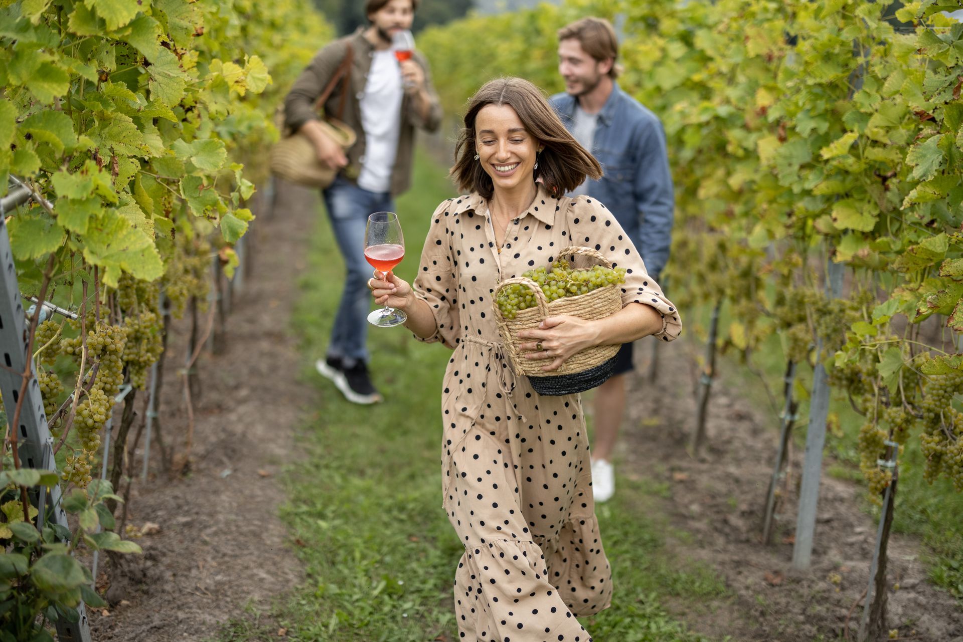 A woman is holding a glass of wine and a basket of grapes in a vineyard.