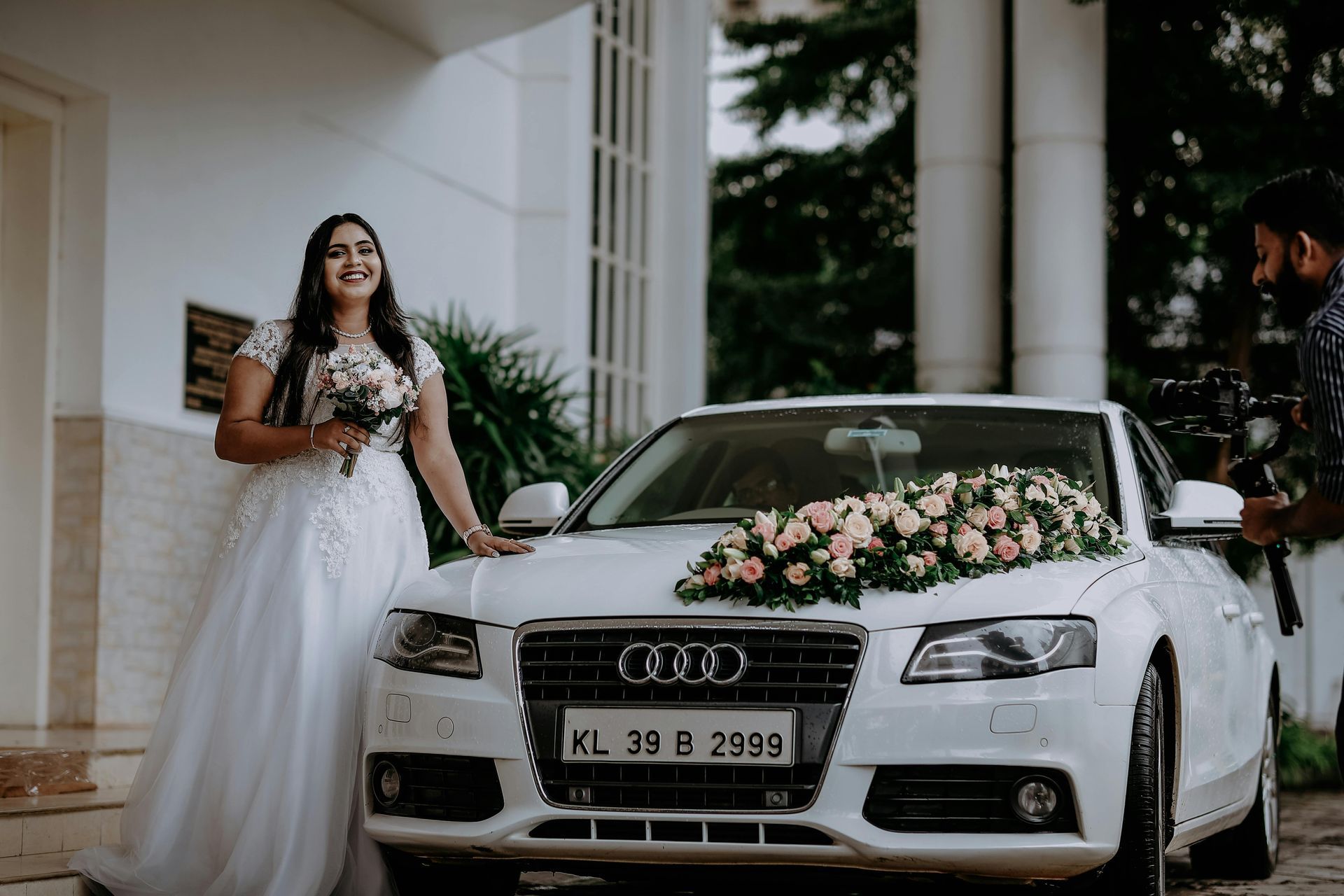 A bride in a white dress is standing next to a white wedding car.