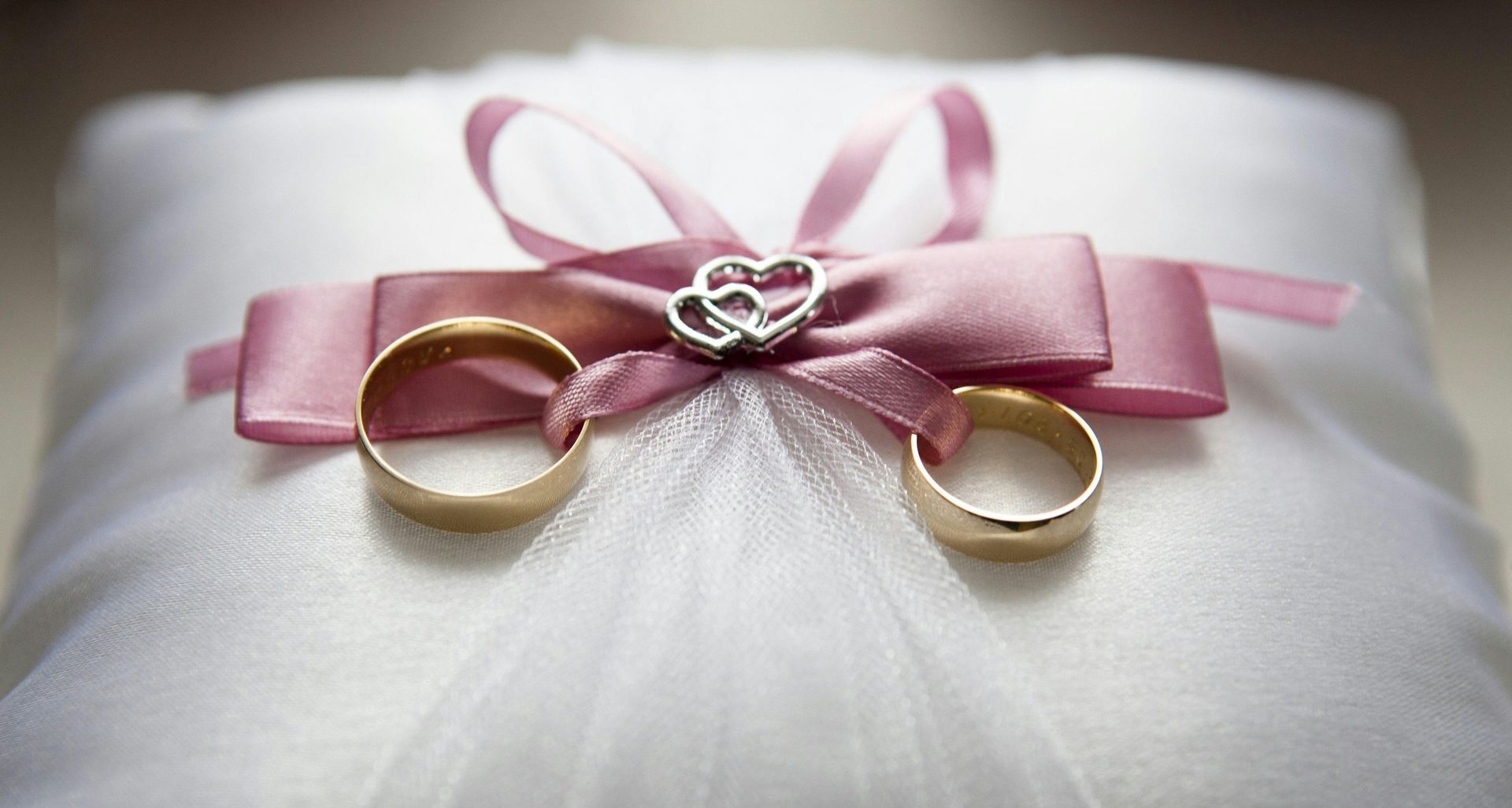 Two wedding rings are sitting on a white pillow with a pink bow.
