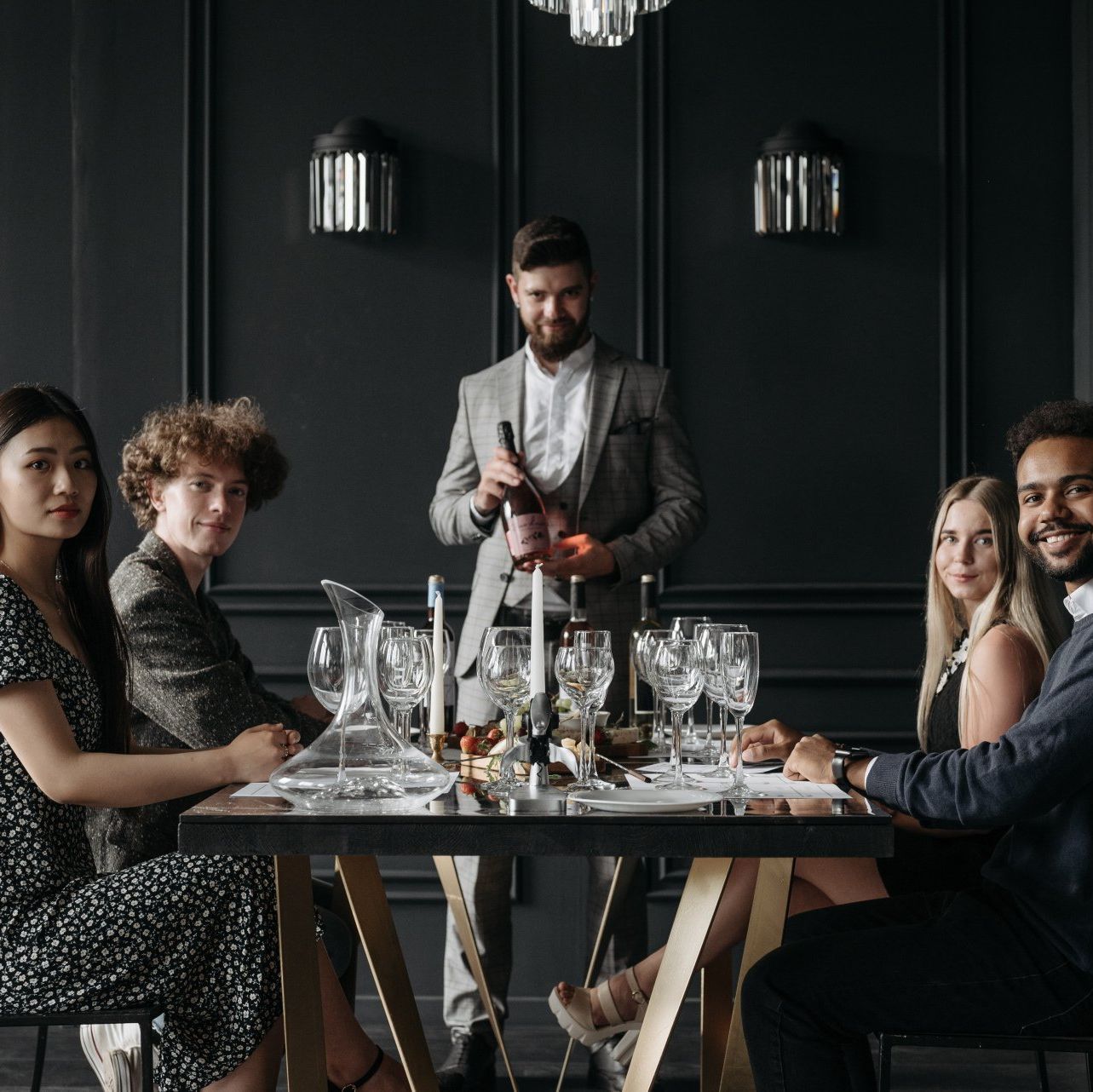 A group of people are sitting at a table with a man standing behind them holding a bottle of wine.