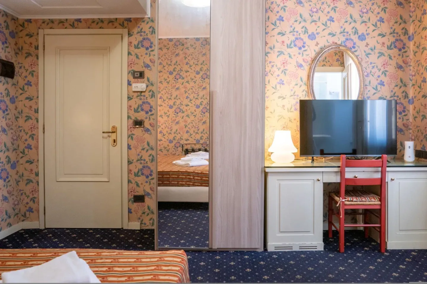 A hotel room with floral wallpaper, a desk with a TV and mirror, and a bed visible in a mirrored section.