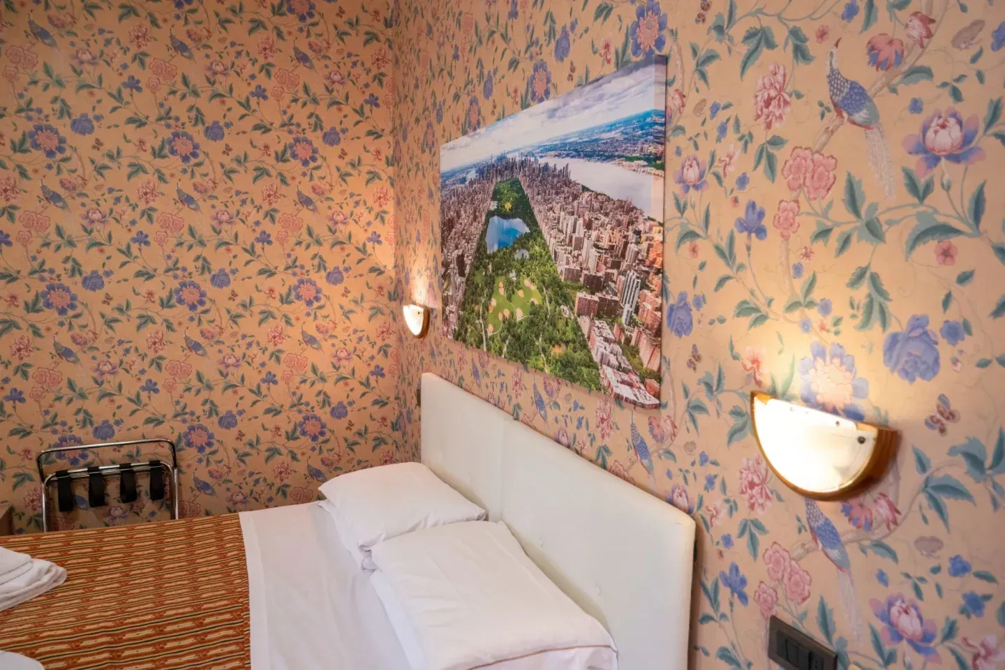 Hotel room with floral wallpaper, a bed with white linens, a New York City aerial view, and sconce lighting.
