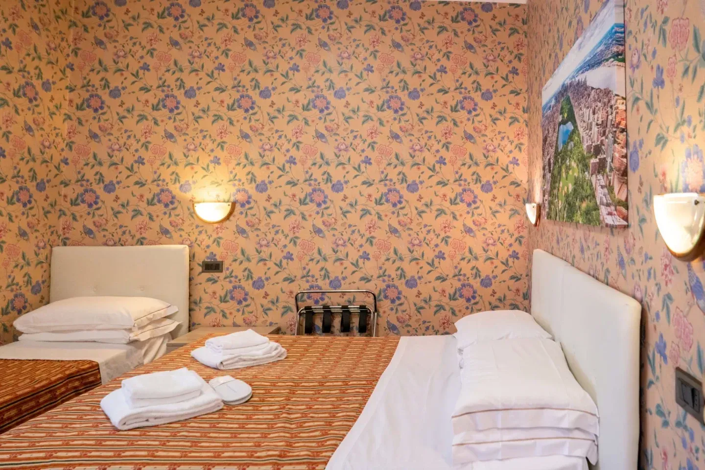 Hotel room with floral wallpaper, two beds, and wall-mounted lights. A photograph hangs on the wall above one bed.