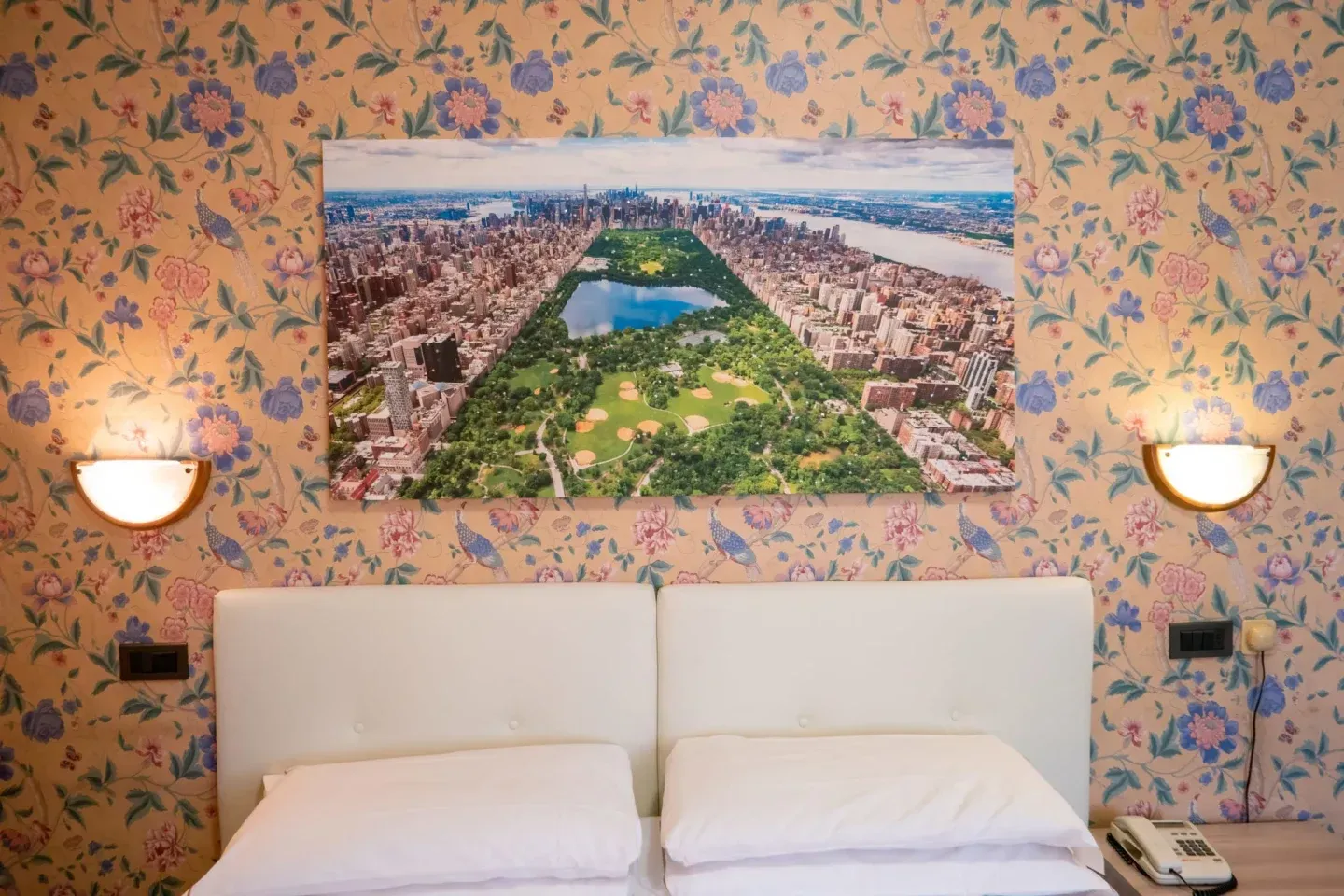 Bedroom with floral wallpaper, a large Central Park photograph, a white headboard, two lamps, and a telephone.