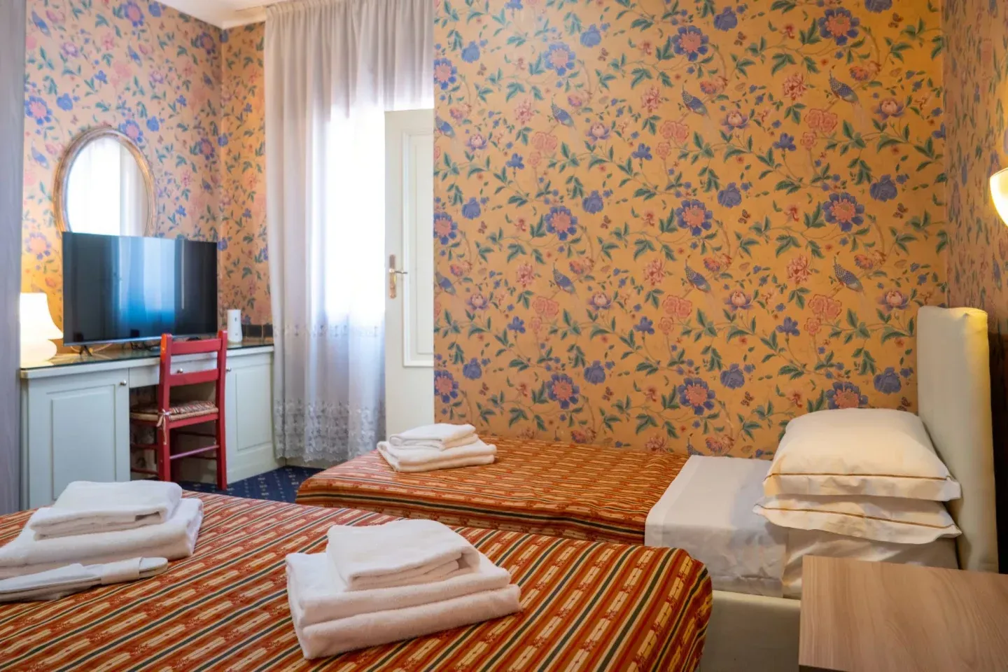 Twin hotel room with patterned wallpaper, beds with folded towels, a TV, and a small desk.