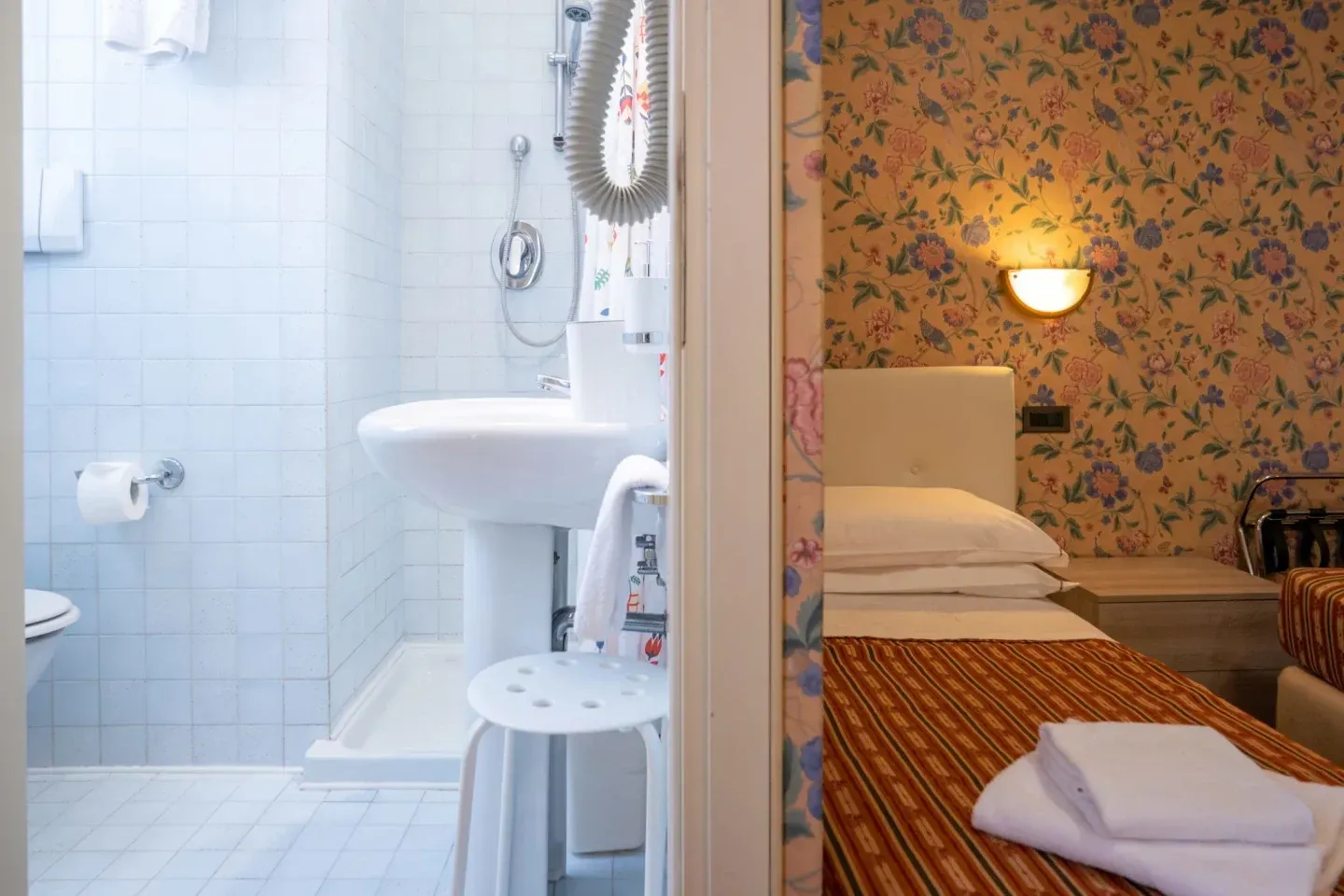 Bathroom with white tile and a sink visible on the left; to the right, a bedroom with floral wallpaper and twin beds.